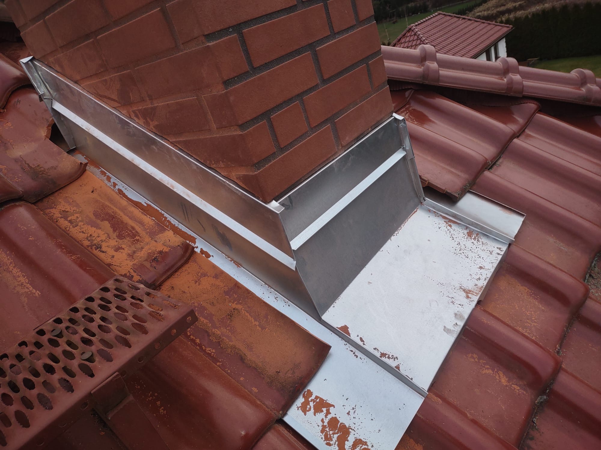 Szczegółowe ujęcie obróbki blacharskiej komina z cegły na dachu pokrytym czerwoną dachówką ceramiczną. Widoczne elementy mocujące i siatka zabezpieczająca.