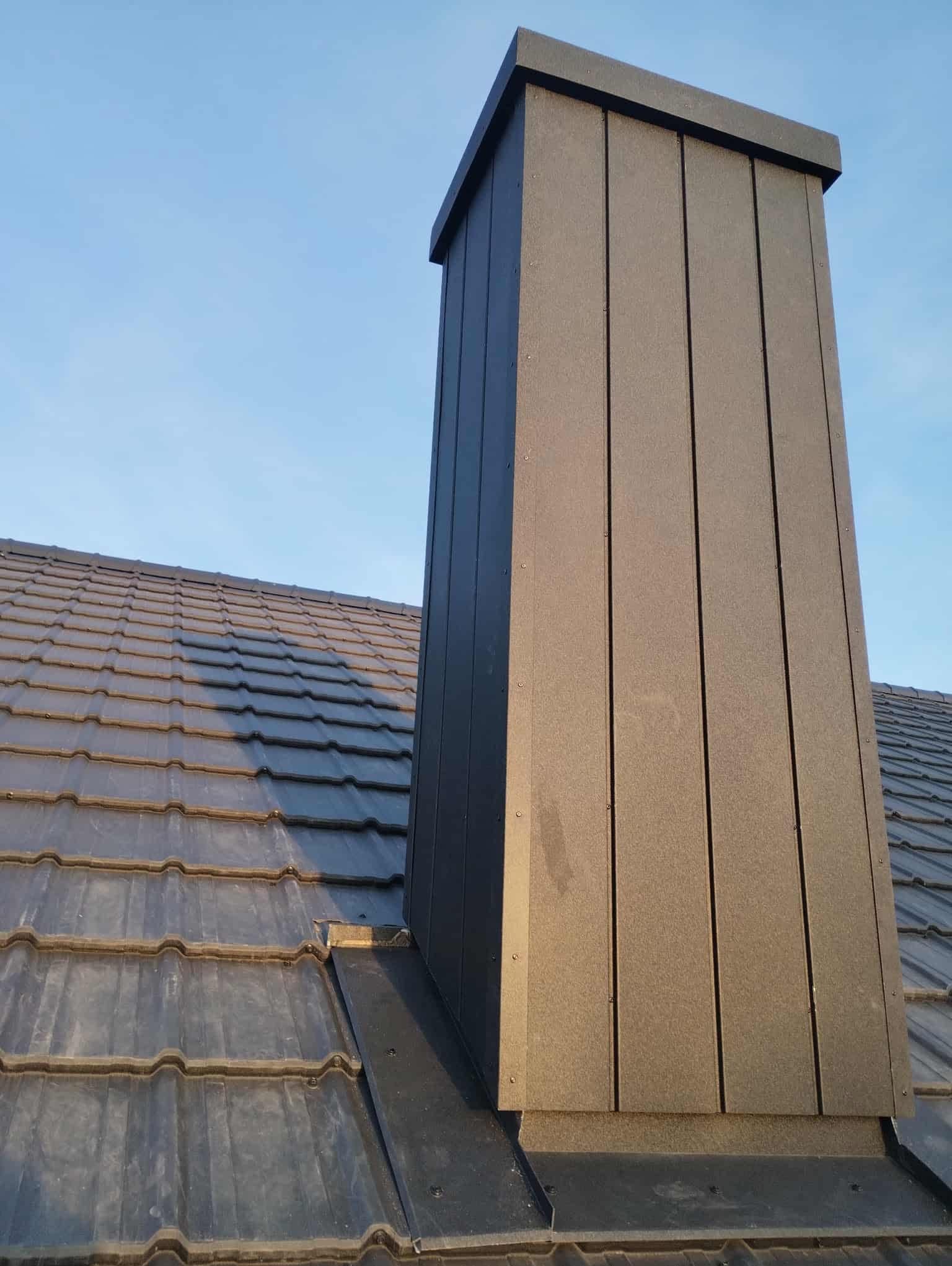 Nowoczesny komin z blachy na dachu z dachówki ceramicznej w Stargardzie. Precyzyjne obróbki blacharskie, estetyczne wykończenie, błękitne niebo w tle.