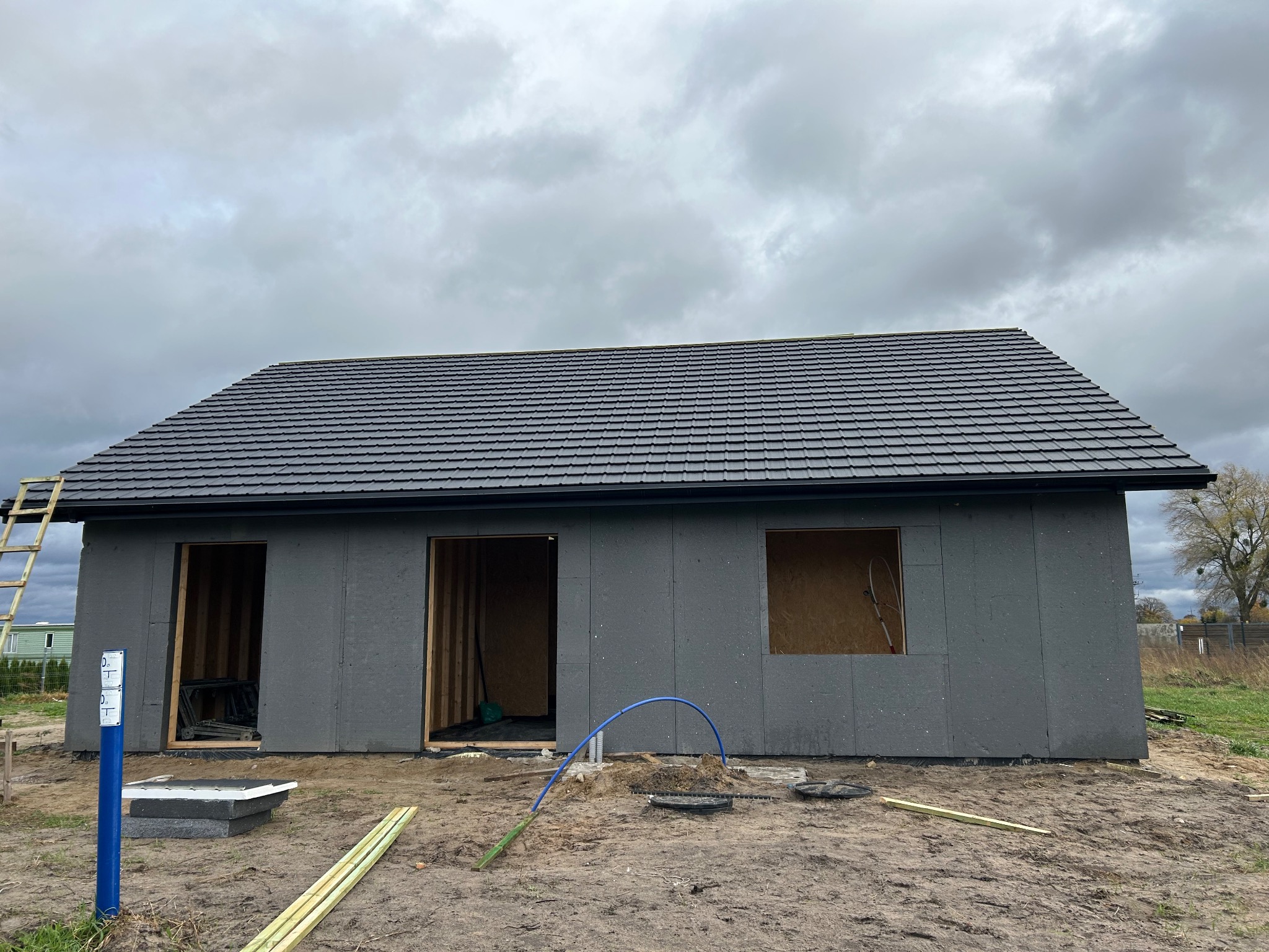 Dom w trakcie budowy z szarymi panelami ściennymi i dachem z ciemnej dachówki. Widoczne otwory okienne i drzwiowe, drabina oparta o ścianę. Pochmurne niebo w tle.