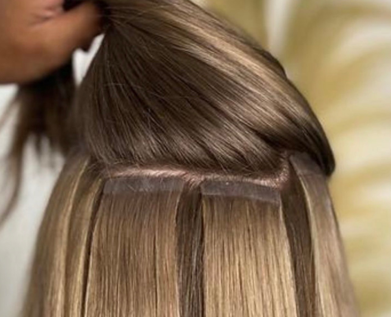 Brązowe włosy z blond pasemkami, przedłużane metodą tape-on. Widoczne łączenia taśmowe, z bliska, w trakcie zabiegu stylizacji.
