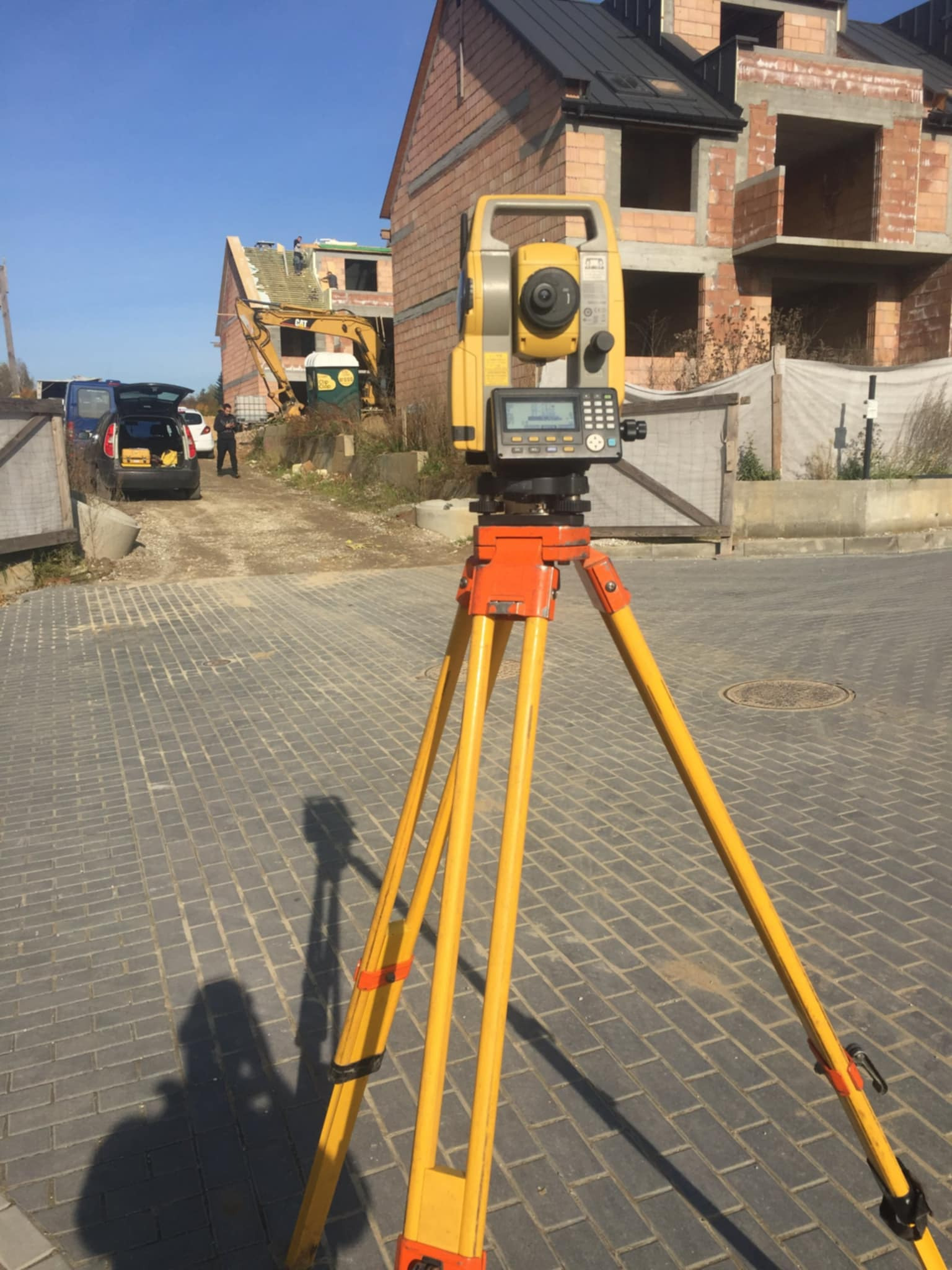 Żółty teodolit na trójnogu na tle placu budowy z widocznym budynkiem w trakcie budowy i koparką. Pomiar geodezyjny w toku.