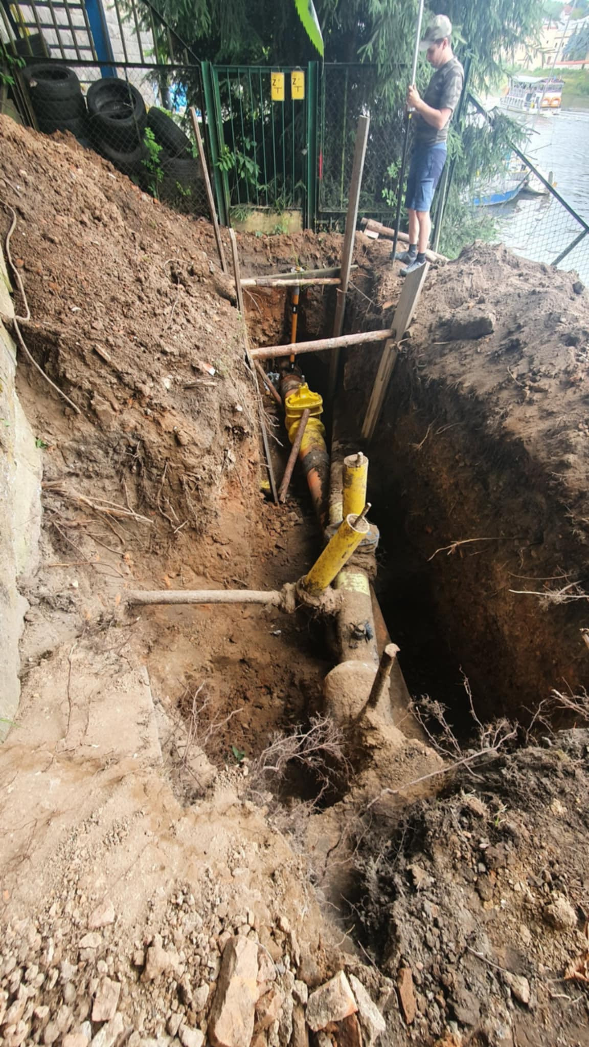 Wykop z widocznymi żółtymi rurami, zabezpieczony drewnianymi belkami. Na górze, przy ogrodzeniu, stoi osoba z tyczką geodezyjną, wykonująca pomiary.