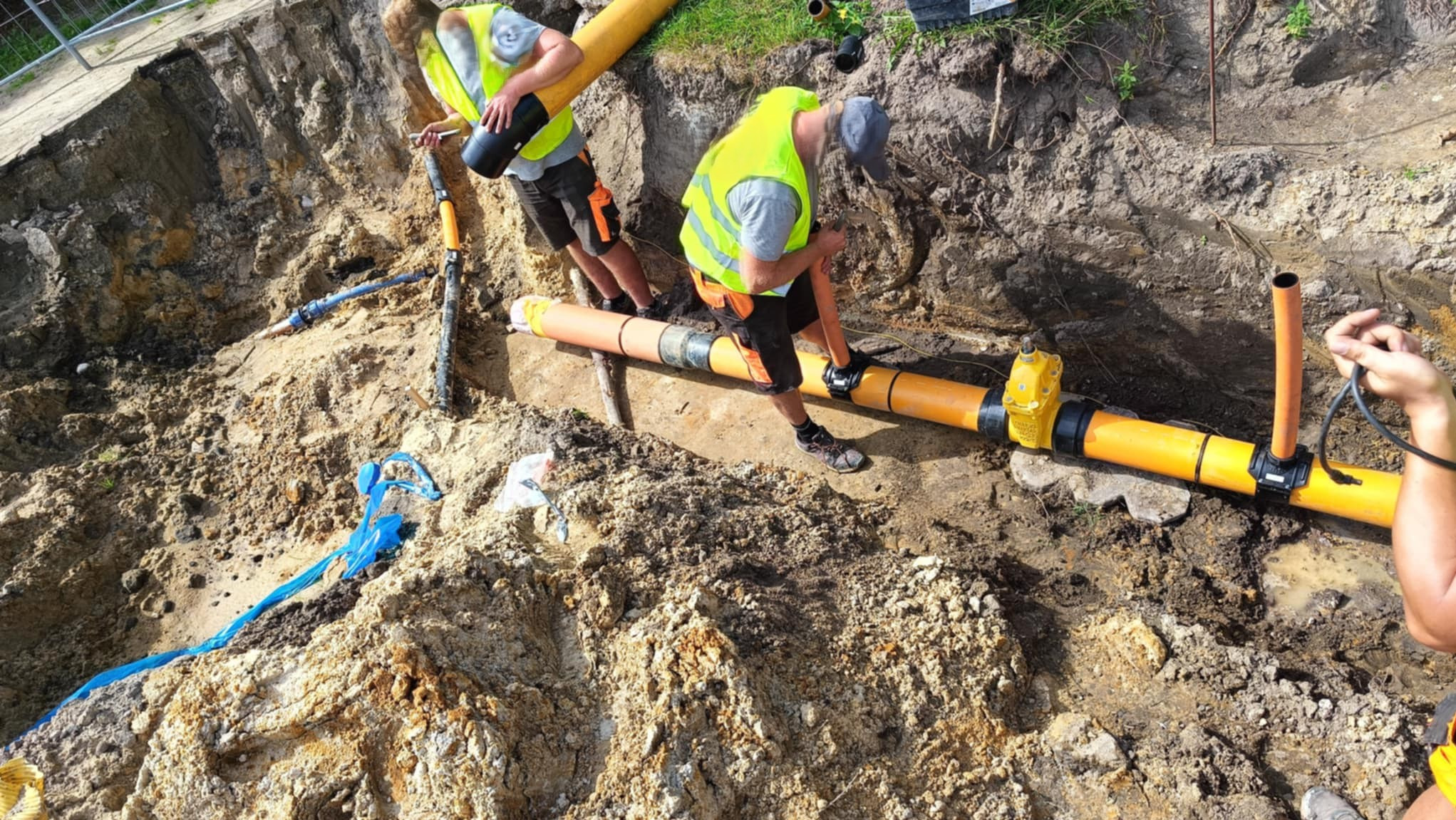 Prace ziemne przy układaniu pomarańczowych rur w wykopie. Dwóch pracowników w kamizelkach odblaskowych łączy rury. Widoczne złącza i fragment wykopu z ziemią.