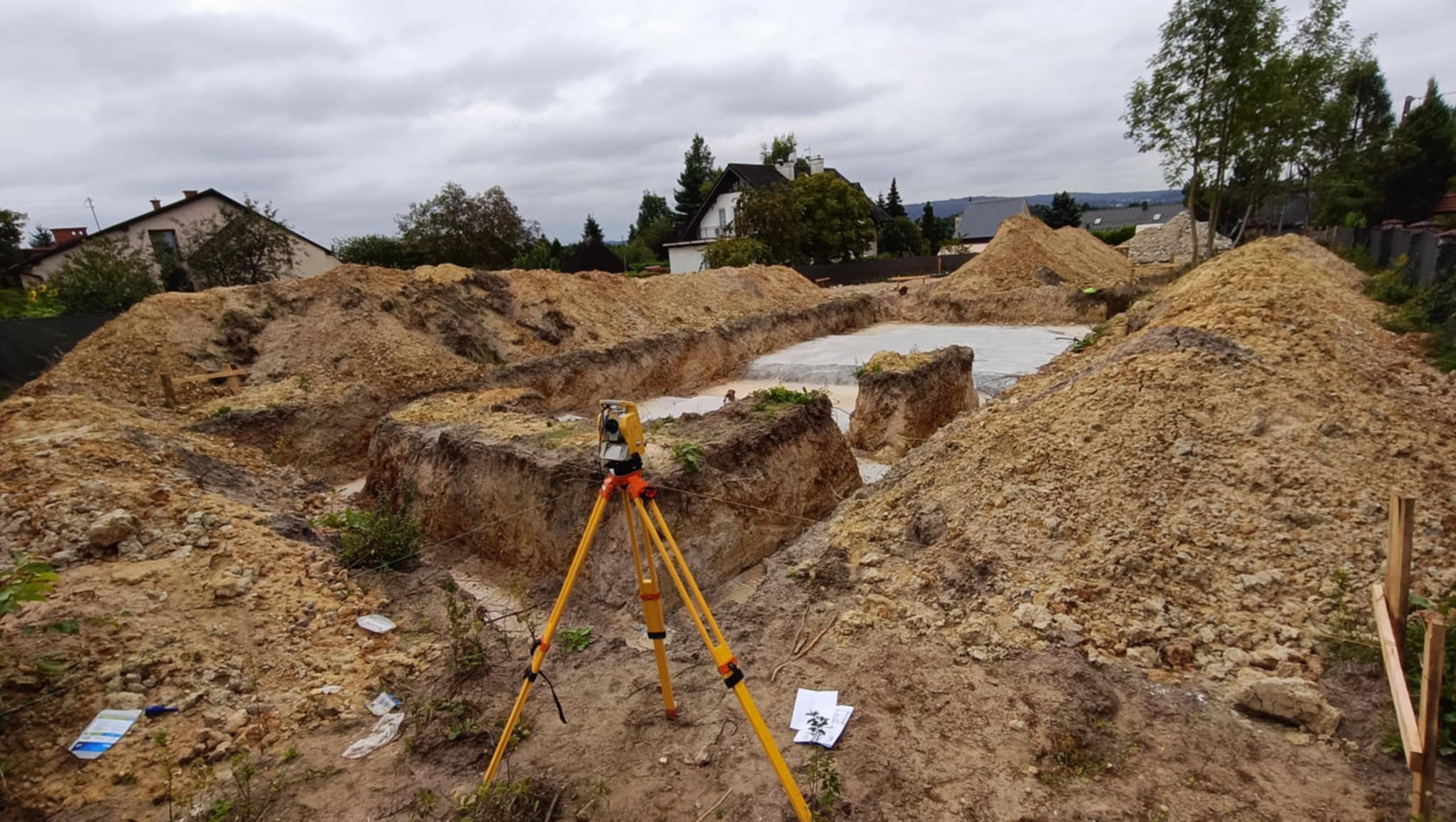 Wytyczanie fundamentów budynku z użyciem niwelatora na statywie. Wykopane rowy fundamentowe, teren budowy z hałdami ziemi. Widok na dom w tle.