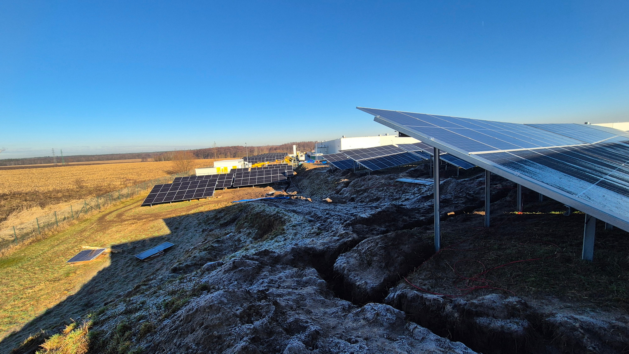 Rozległa instalacja paneli słonecznych na nierównym terenie, z widocznymi śladami prac ziemnych i budynkiem w tle, uchwycona w jasnym, słonecznym świetle.