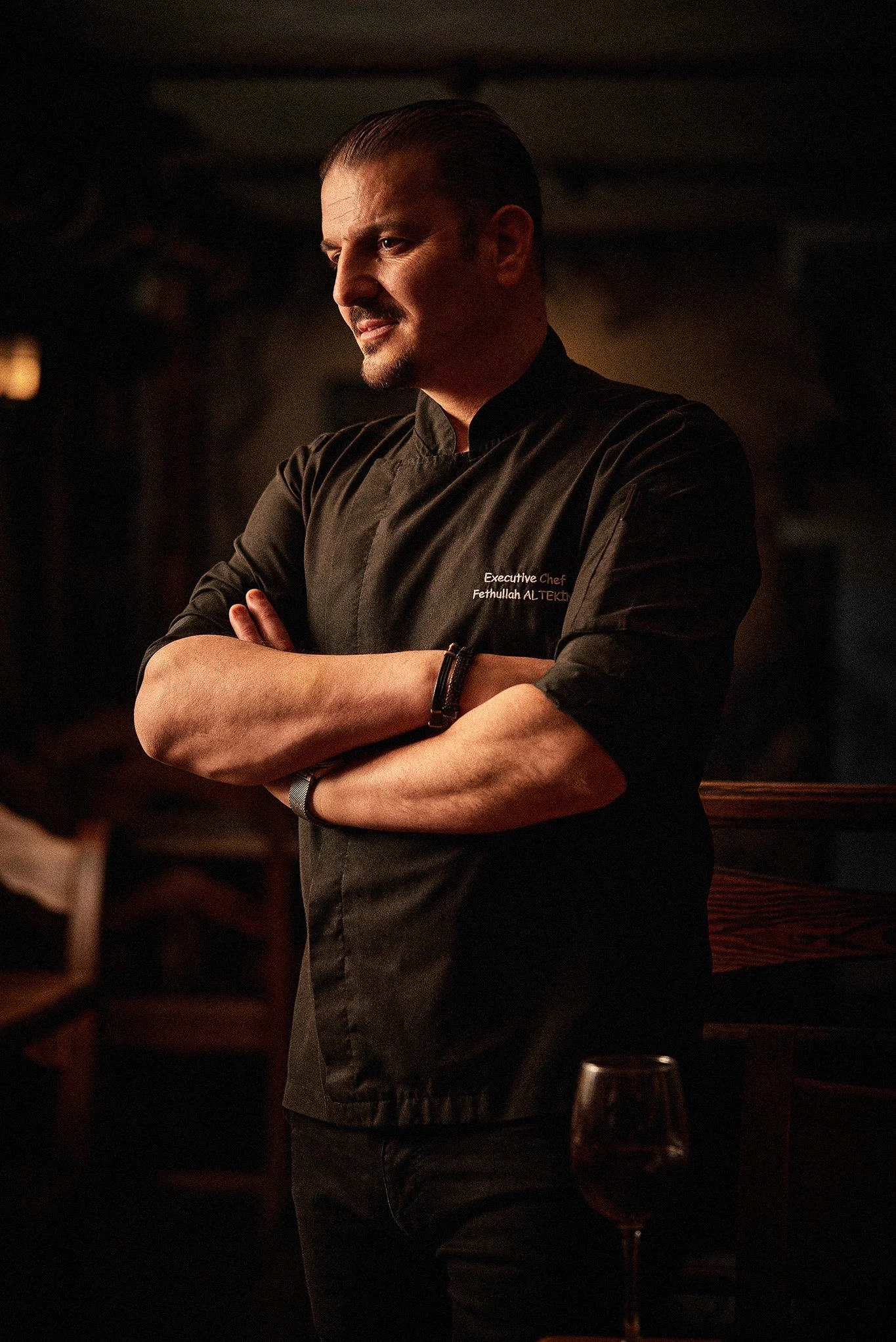 Portret mężczyzny w czarnym uniformie szefa kuchni z wyhaftowanym imieniem i nazwiskiem na tle ciemnego wnętrza restauracji z kieliszkiem wina.