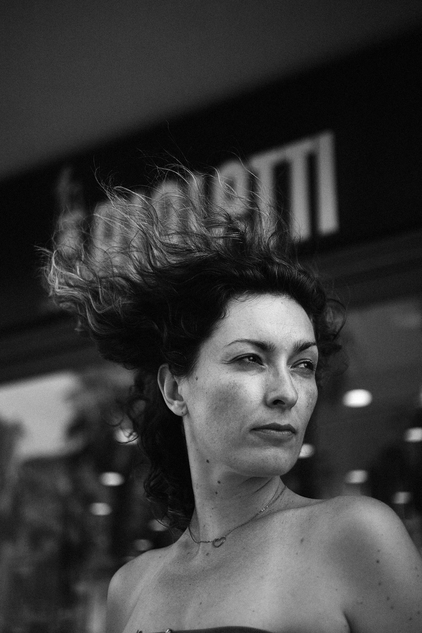 Czarno-biały portret kobiety z rozwianymi włosami, spoglądającej w prawo, z widoczną delikatną biżuterią na szyi. Tło rozmyte, z elementami architektonicznymi.