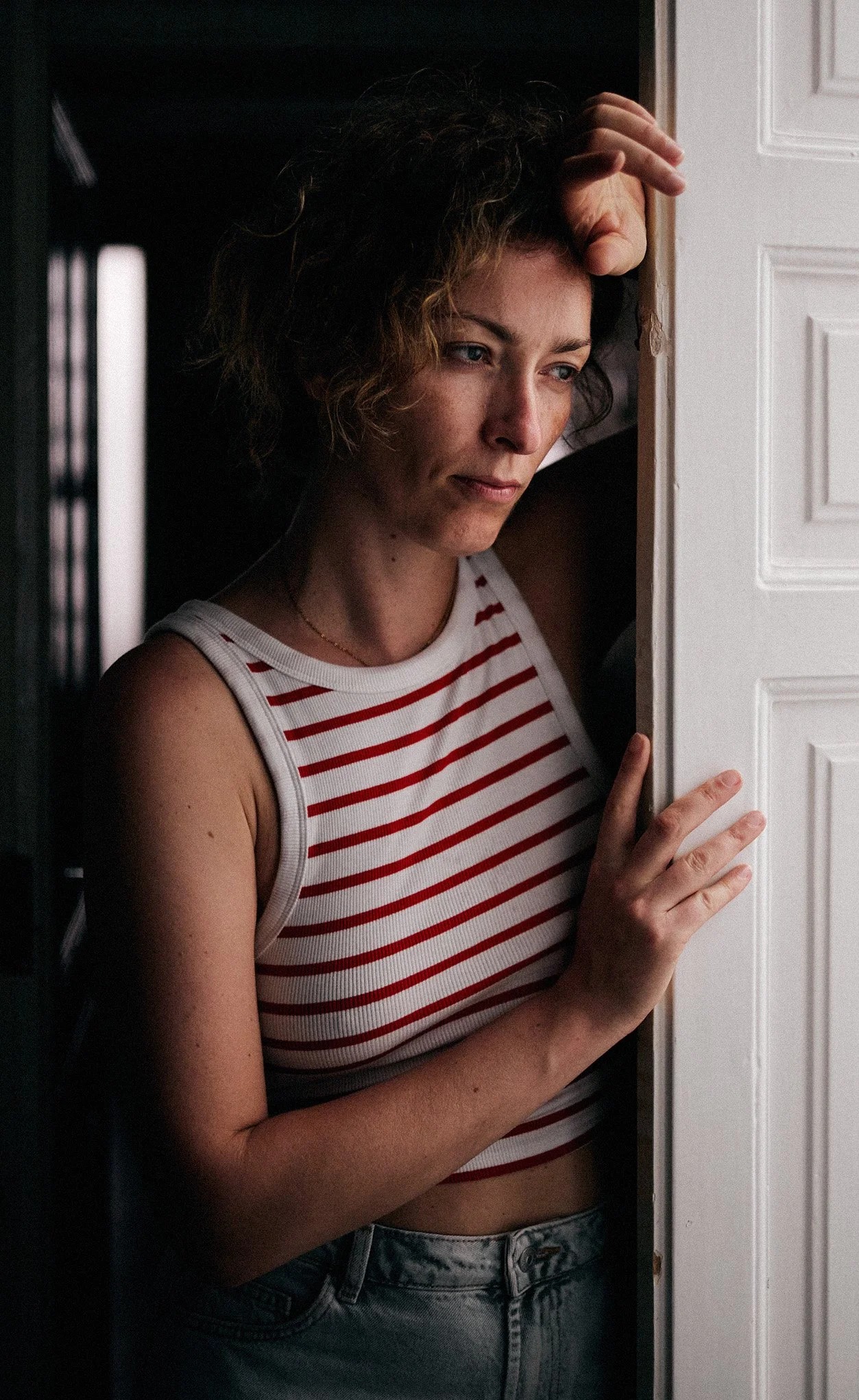 Portret kobiety w paski, opierającej się o białe drzwi, zamyślona twarz, miękkie światło podkreśla rysy, ciemne tło dodaje głębi. Kadrowanie od pasa w górę.