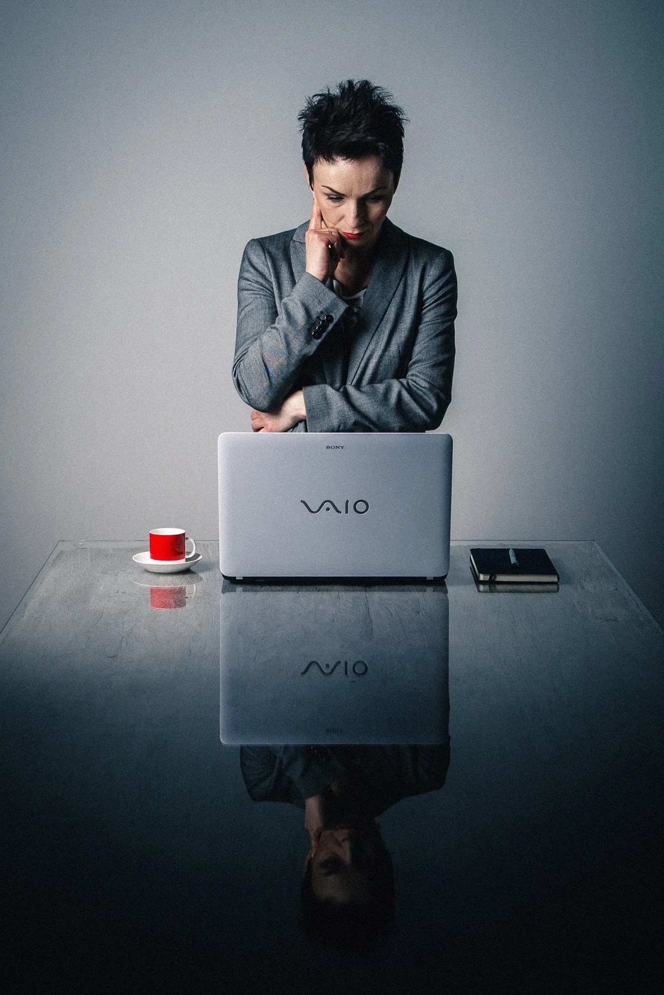 Kobieta w szarym żakiecie zamyślona przed laptopem Sony Vaio, obok czerwona filiżanka i notatnik na szklanym biurku z odbiciem. Portret biznesowy w chłodnej tonacji.