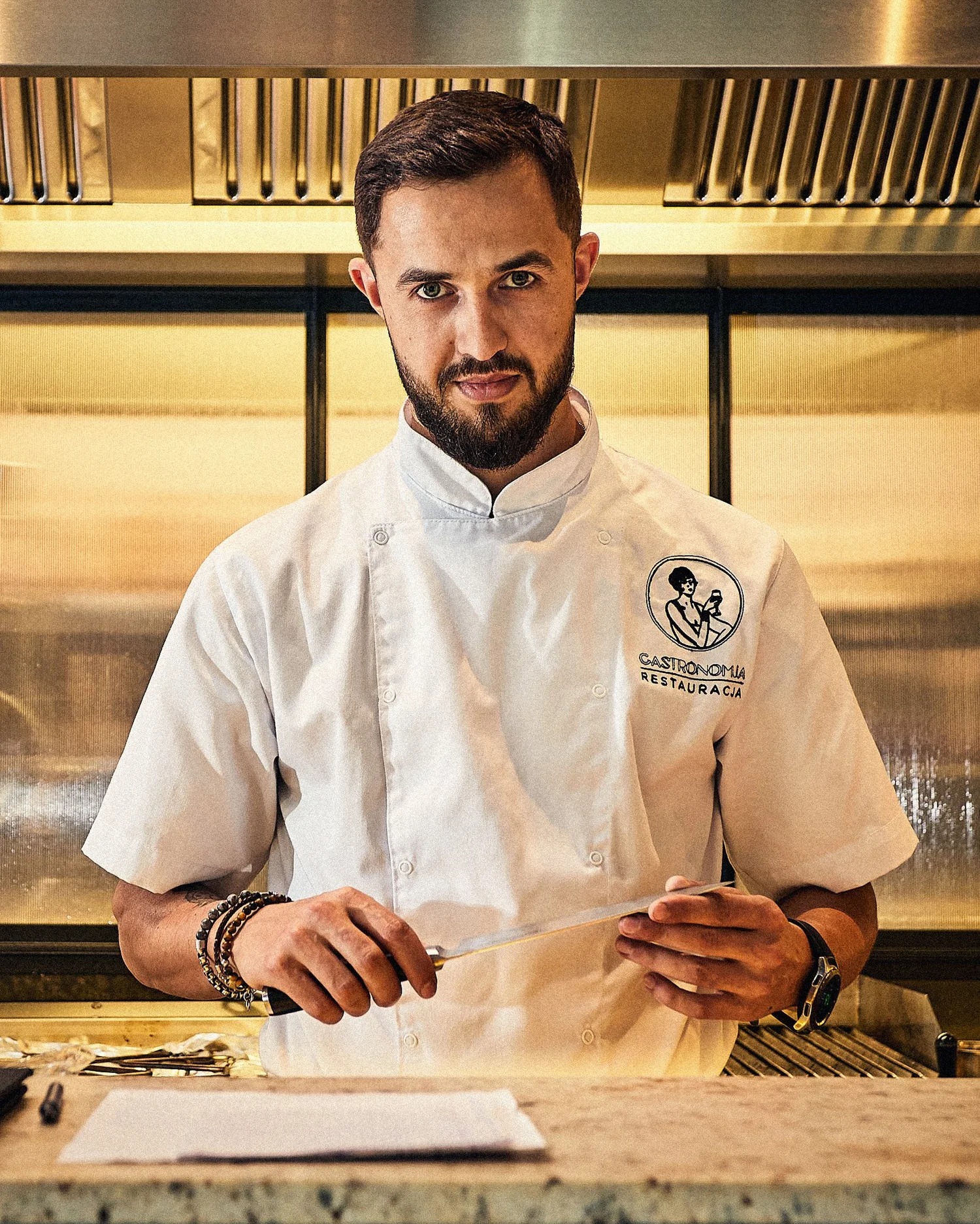 Portret kucharza w białym uniformie z logo 'Gastronomia Restauracja', trzymającego nóż na tle profesjonalnej kuchni z metalowymi elementami i ciepłym oświetleniem.