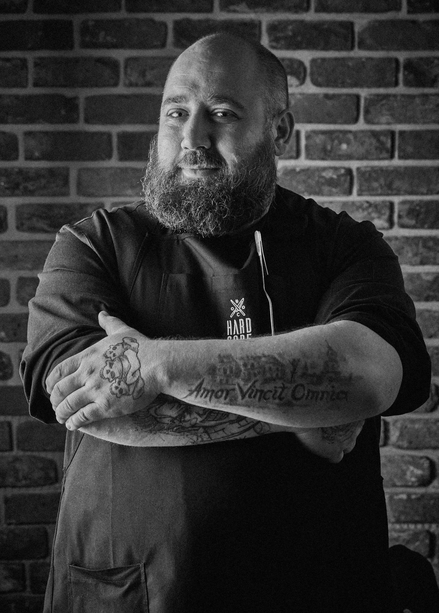 Czarno-biały portret brodatego mężczyzny z tatuażami na tle ceglanej ściany, ubrany w fartuch z logo 'HARD CODE'.