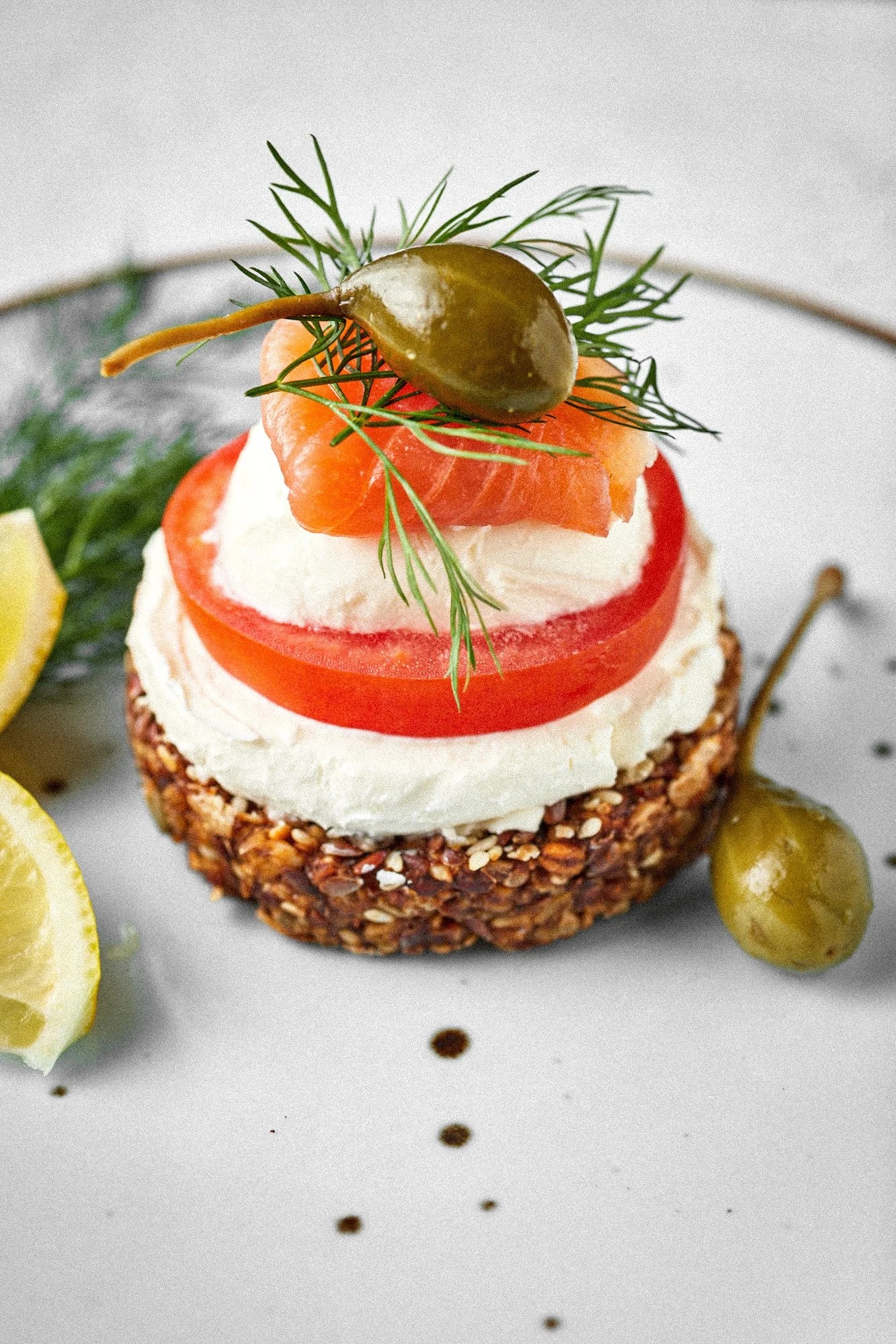 Apetyczna kanapka z łososiem, kremowym serem, pomidorem i kaparami, udekorowana koperkiem, na talerzu z kroplami sosu i cytryną. Delikatne światło, bliski kadr.