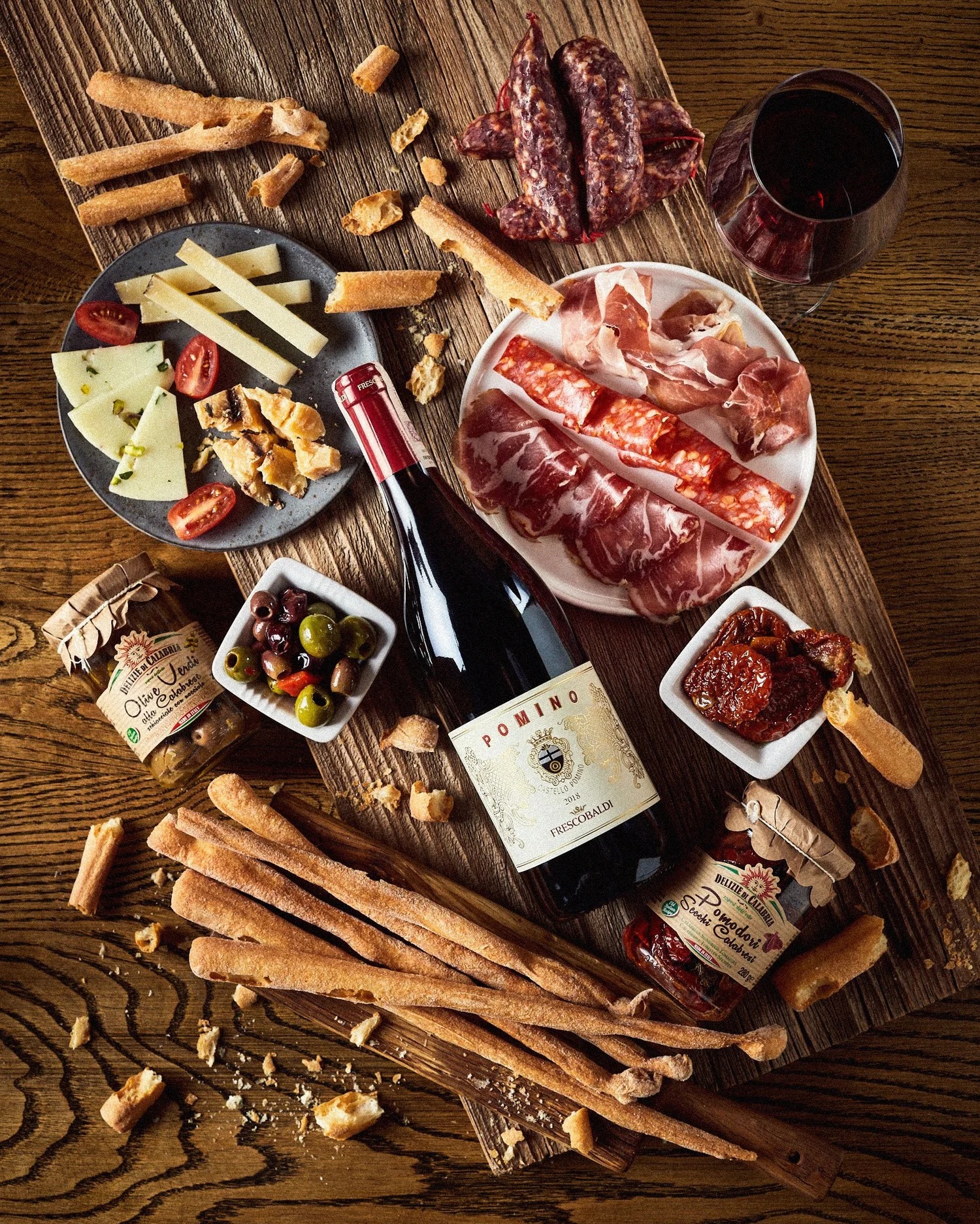Deska serów i wędlin z oliwkami, pomidorami suszonymi, grissini i butelką czerwonego wina Pomino Frescobaldi na drewnianym stole z wyraźną fakturą.