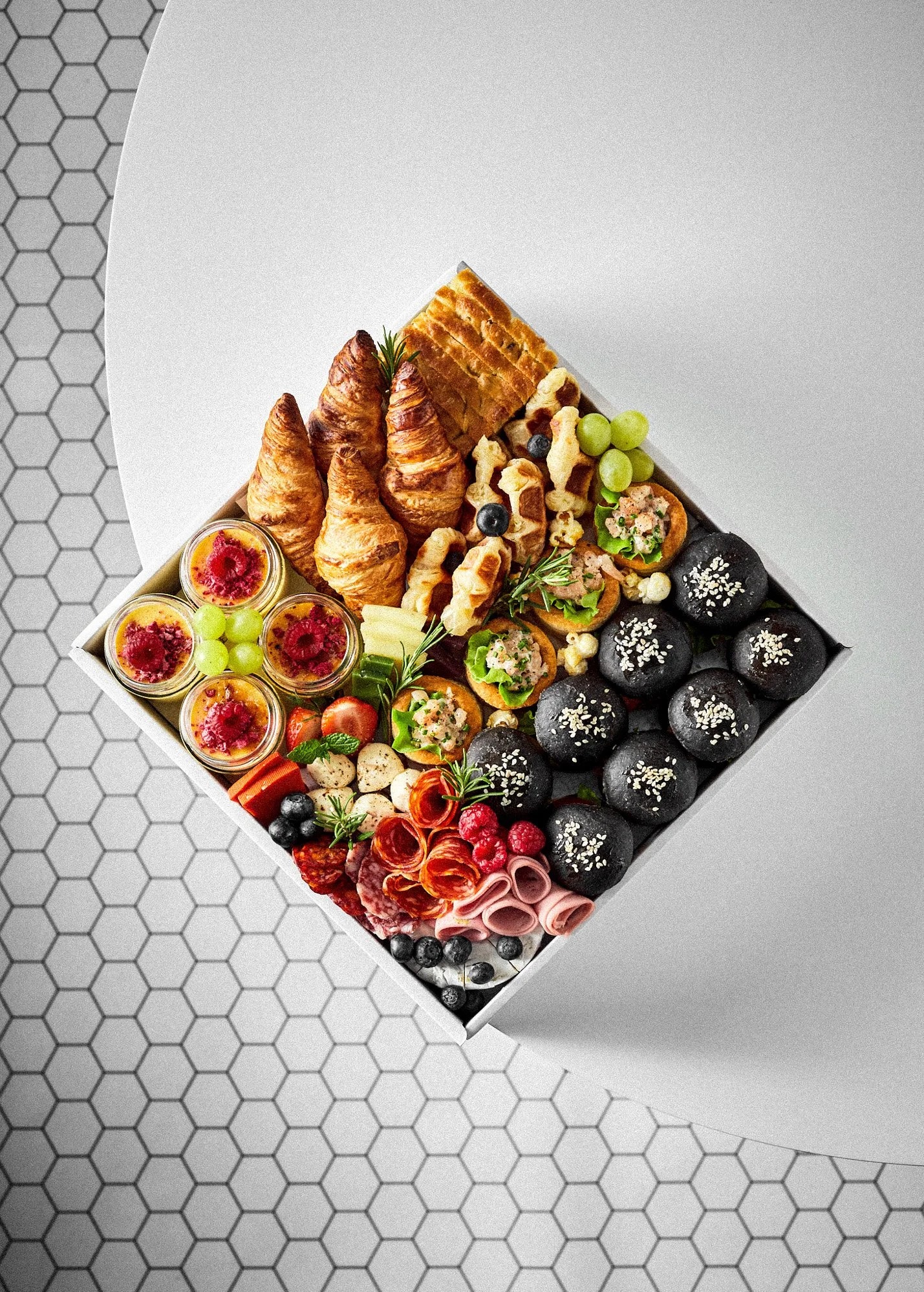 Elegancka kompozycja kulinarna w pudełku: croissanty, gofry, desery w słoiczkach, mini-kanapki i ciemne bułeczki z sezamem. Widok z góry na tle białego stołu i heksagonalnej podłogi.