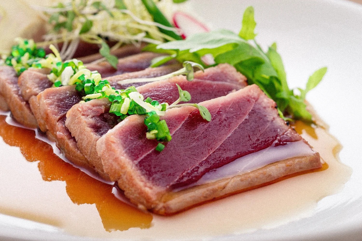 Selekcja tataki z tuńczyka, udekorowana świeżymi ziołami i skropiona sosem, na białym talerzu. Delikatne, miękkie światło podkreśla strukturę ryby.