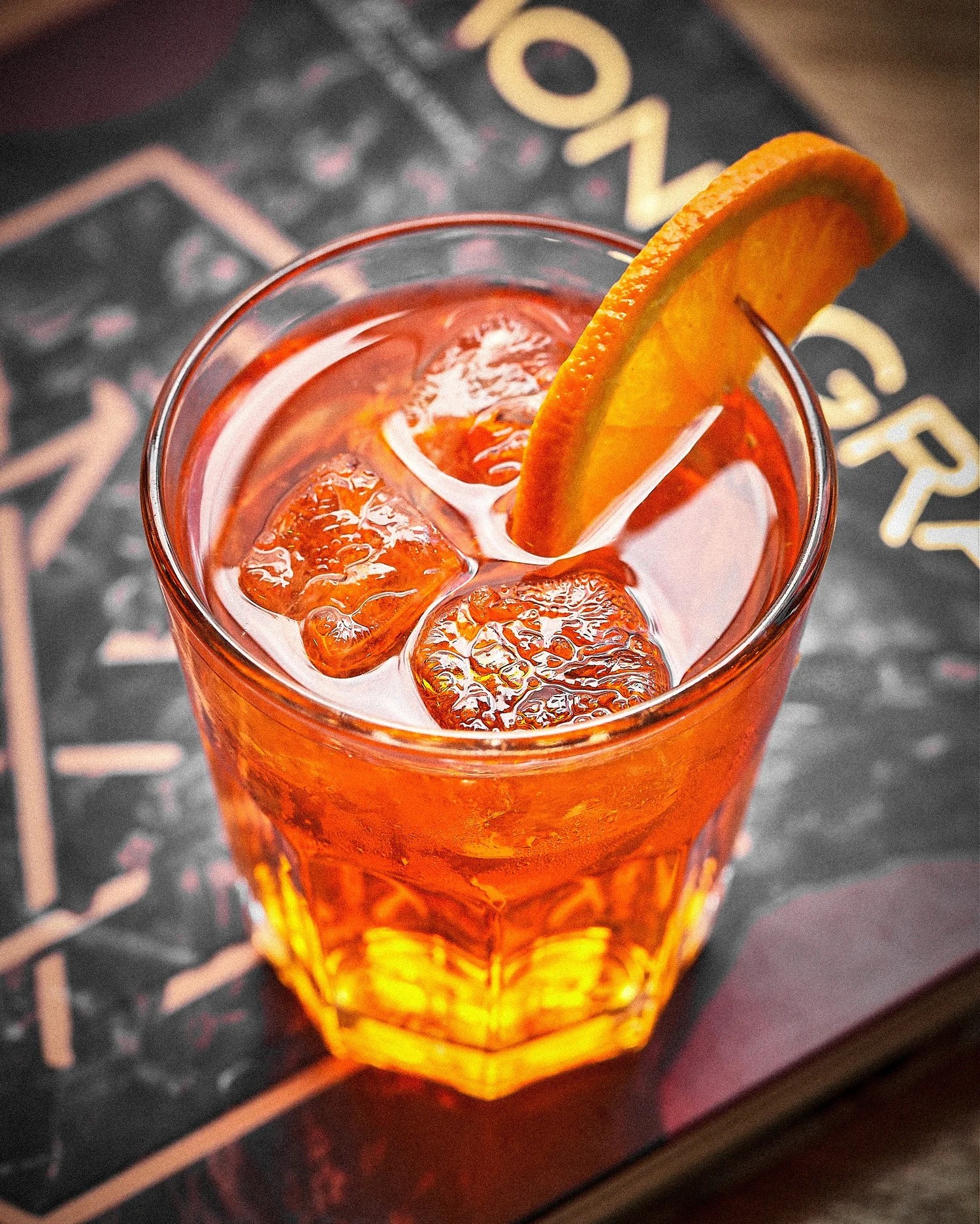 Kadr z góry na szklankę Aperol Spritz z lodem i pomarańczą, ustawioną na książce z geometrycznym wzorem. Ciepłe światło podkreśla pomarańczowy kolor napoju.
