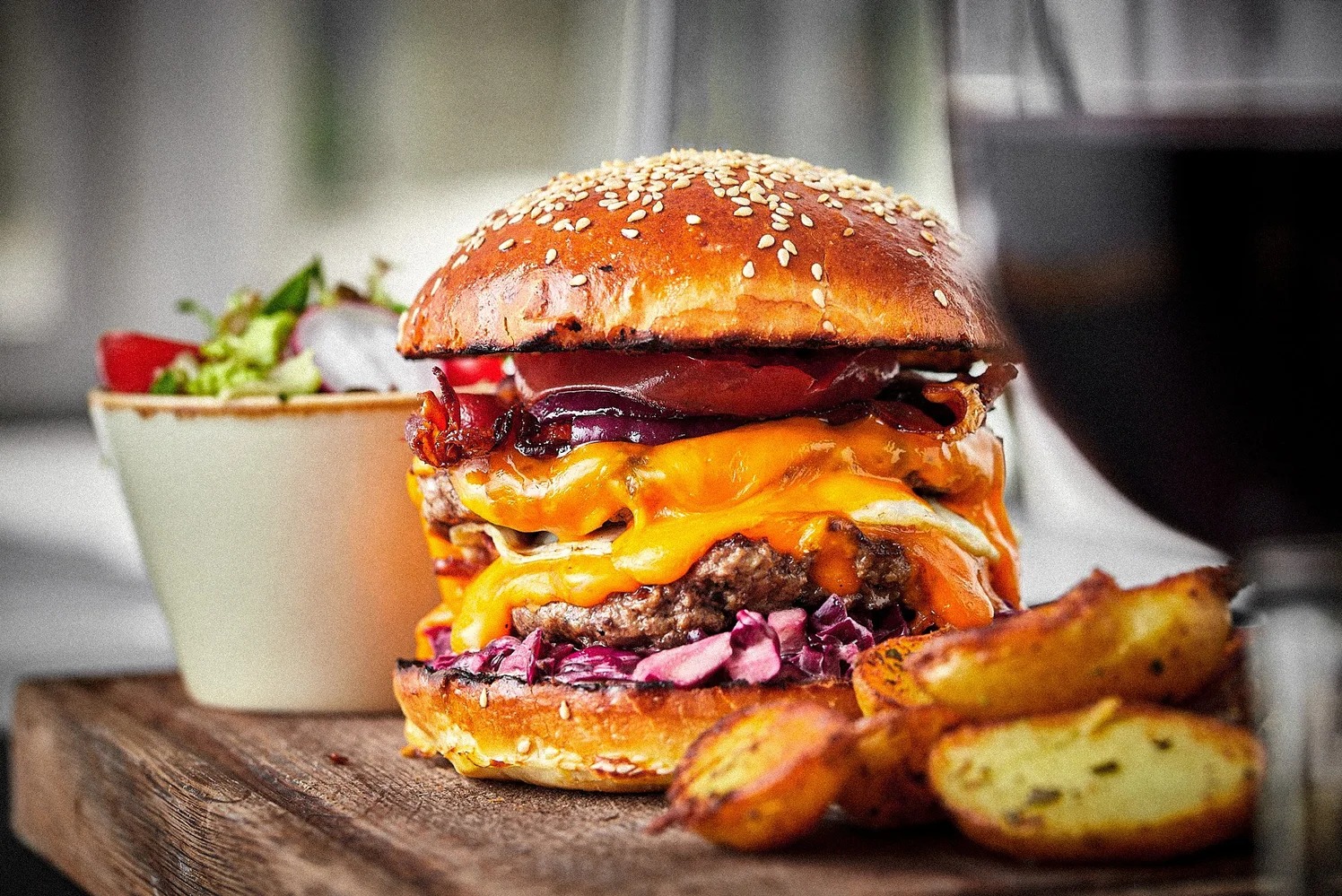 Apetyczny, dwuwarstwowy burger z serem cheddar, bekonem i czerwoną cebulą, podany na drewnianej desce z sałatką i ziemniakami, w otoczeniu jasnego tła.
