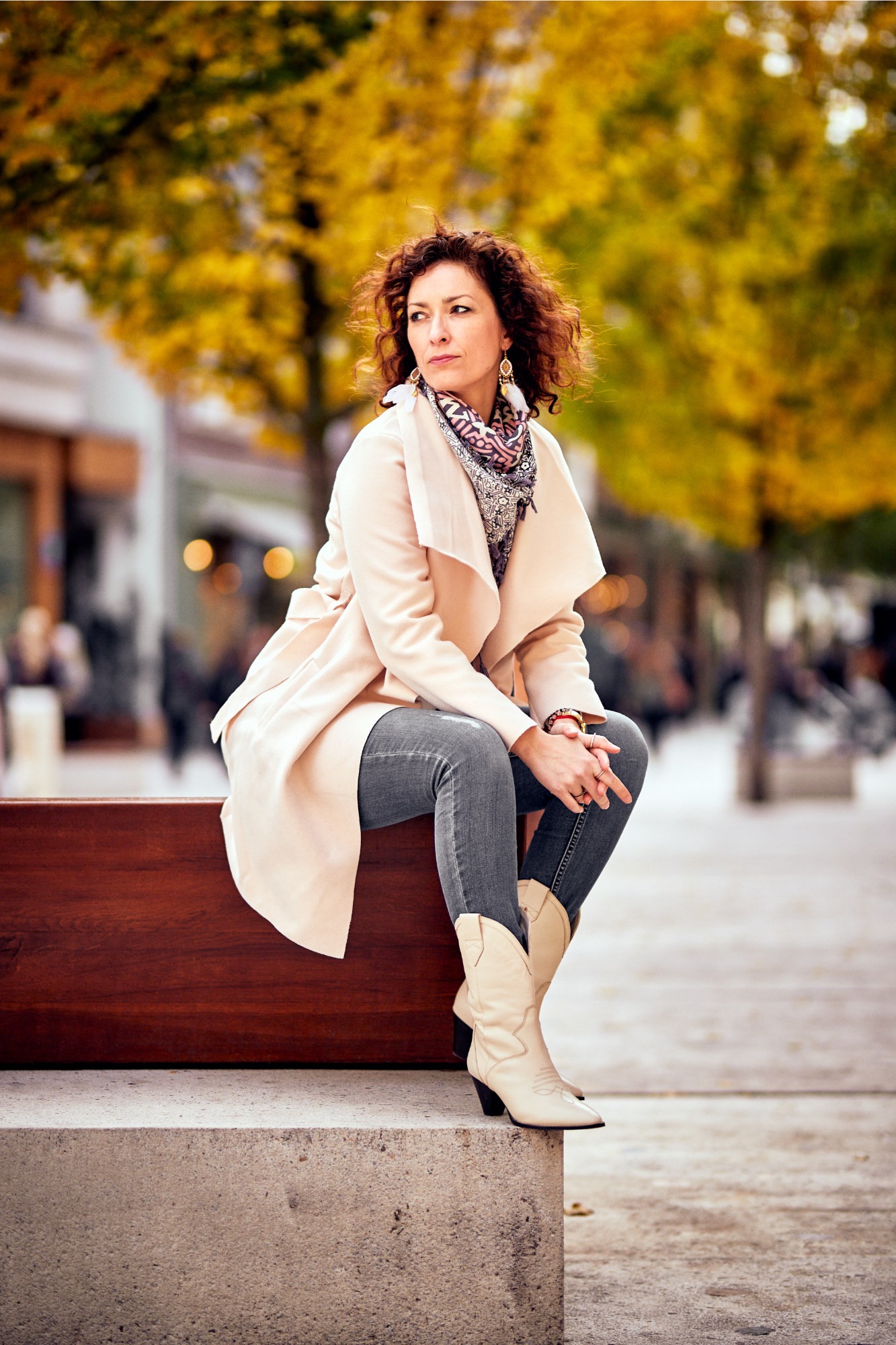 Kobieta w płaszczu i kowbojkach siedzi na ławce w parku jesienią, spoglądając w bok. Uwagę przykuwają kręcone włosy i wzorzysta apaszka.