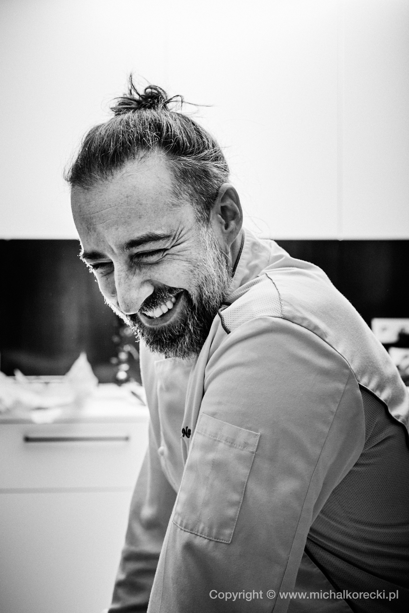 Czarno-biały portret uśmiechniętego mężczyzny z brodą i włosami spiętymi w kok, w uniformie, uchwycony w naturalnym świetle w Warszawie. Wyraz radości.