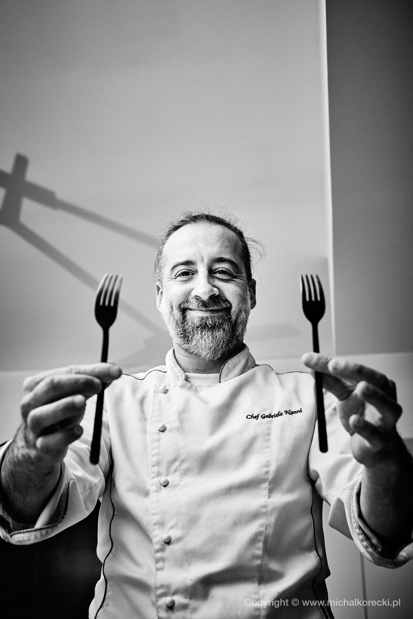 Czarno-białe zdjęcie uśmiechniętego szefa kuchni Gabriele Nonni trzymającego dwa widelce, z widocznym logo fotografa w dolnej części kadru.