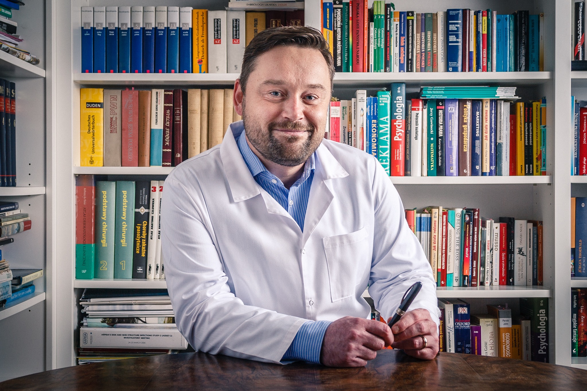 Lekarz w białym kitlu siedzi za biurkiem, trzymając pióro. W tle regał z książkami. Portret w gabinecie lekarskim, ciepłe oświetlenie, profesjonalny wizerunek.