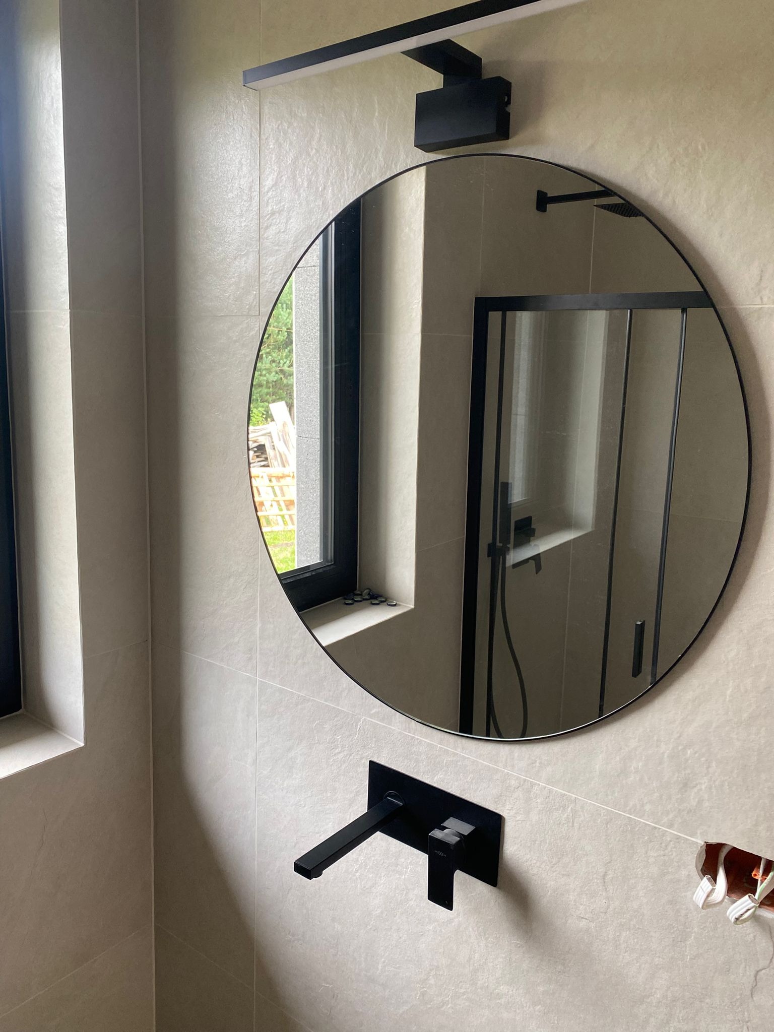 Minimalistyczna łazienka z okrągłym lustrem i czarną armaturą. W lustrze odbija się kabina prysznicowa i okno z zielenią. Widoczne płytki w odcieniach beżu.