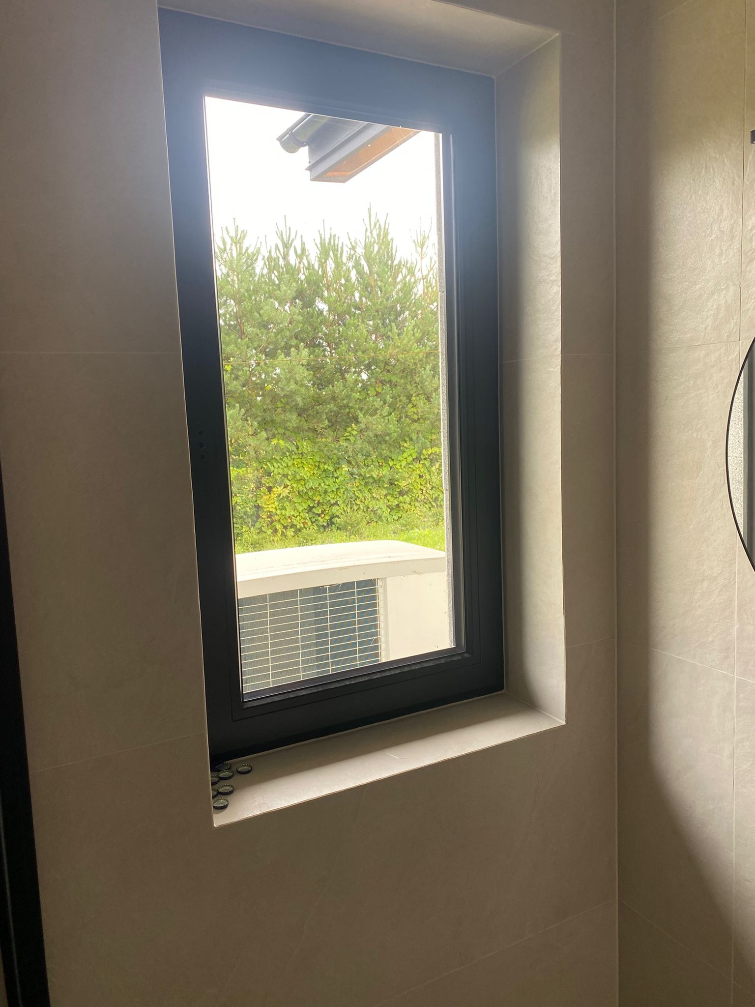 Wnętrze łazienki w Toruniu z oknem, przez które widać zieleń. Ściany wyłożone jasnymi płytkami. Na parapecie leżą kapsle od piwa.