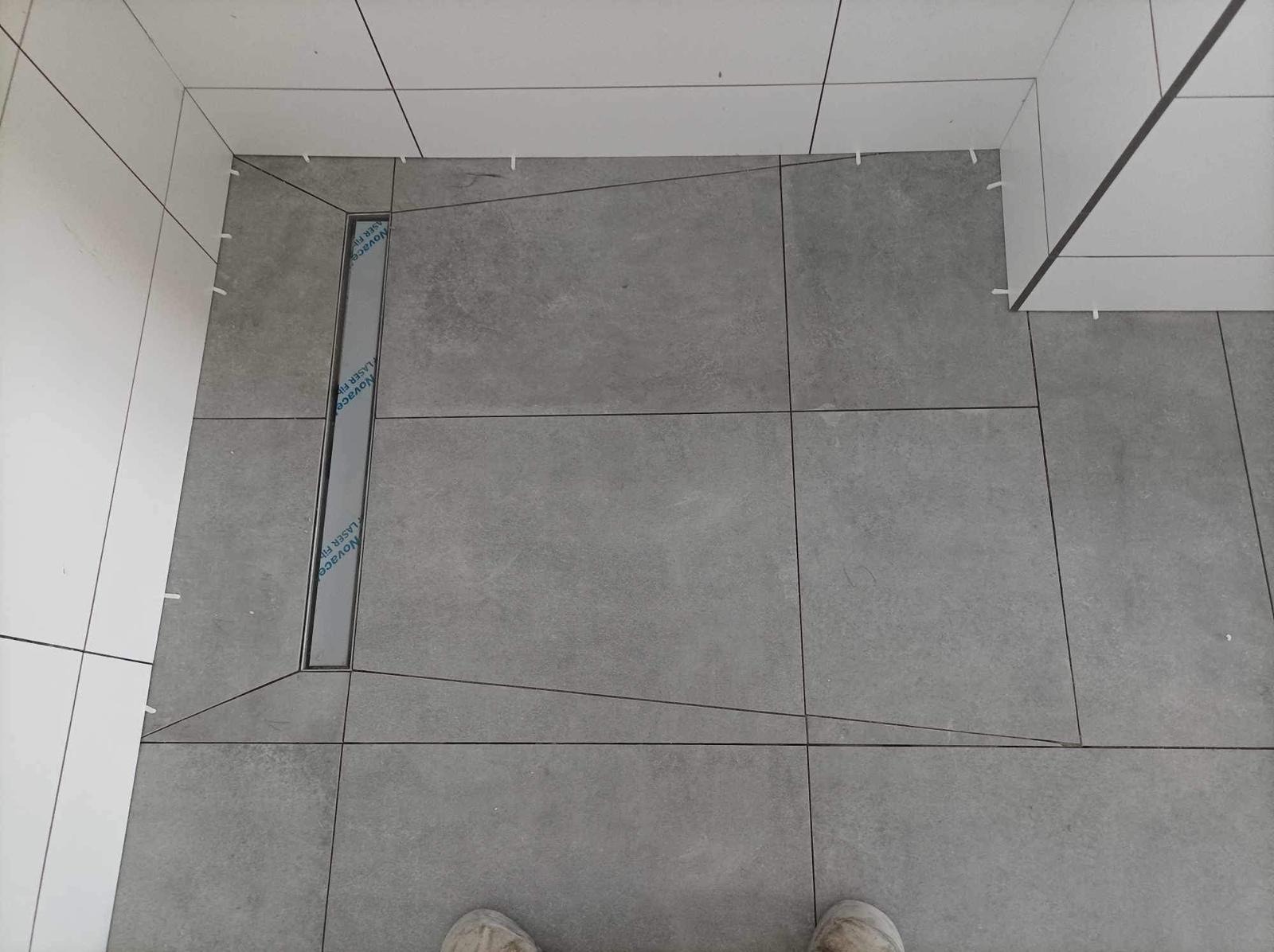 Ułożone szare płytki podłogowe z odpływem liniowym w kabinie prysznicowej. Widoczne białe płytki na ścianach i fragment butów w dolnej części kadru.