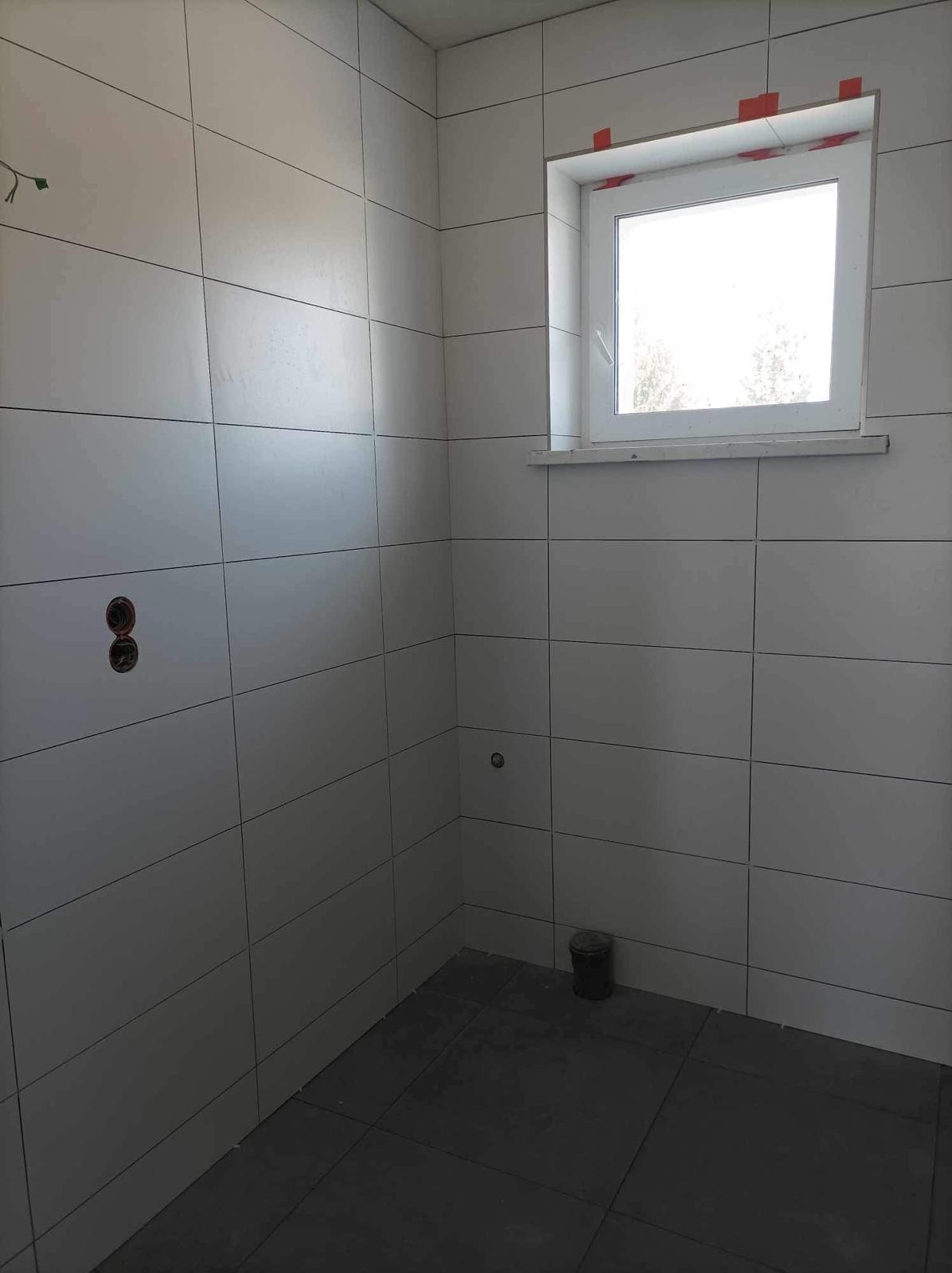 Wykończona łazienka z białymi, prostokątnymi płytkami na ścianach i ciemnymi płytkami na podłodze. Widoczne gniazdka elektryczne i okno z taśmą zabezpieczającą.