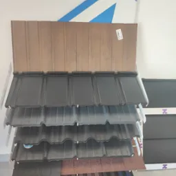 Ekspozycja różnorodnych pokryć dachowych na stojaku w sklepie, prezentacja kolorów i faktur blachodachówki, z widocznym logo producenta na tle białej ściany.