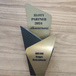 Nagroda 'Złoty Partner 2024' dla firmy BLACHOTRAPEZ oraz Greg-Bud Grzegorz Siestrzewitowski, na drewnianym tle. Trapezoidalny kształt, złote i czarne elementy.
