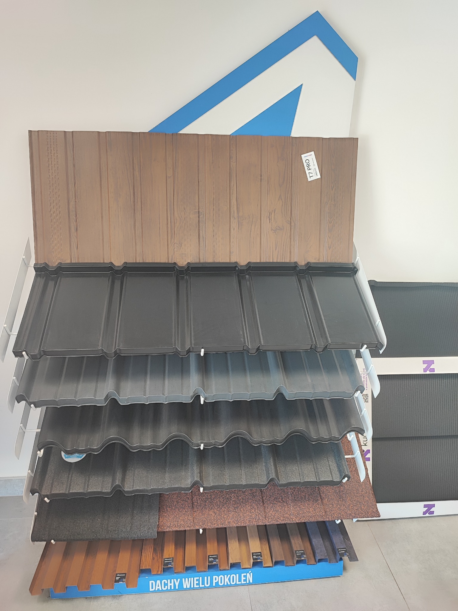 Ekspozycja różnorodnych pokryć dachowych na stojaku w sklepie, prezentacja kolorów i faktur blachodachówki, z widocznym logo producenta na tle białej ściany.