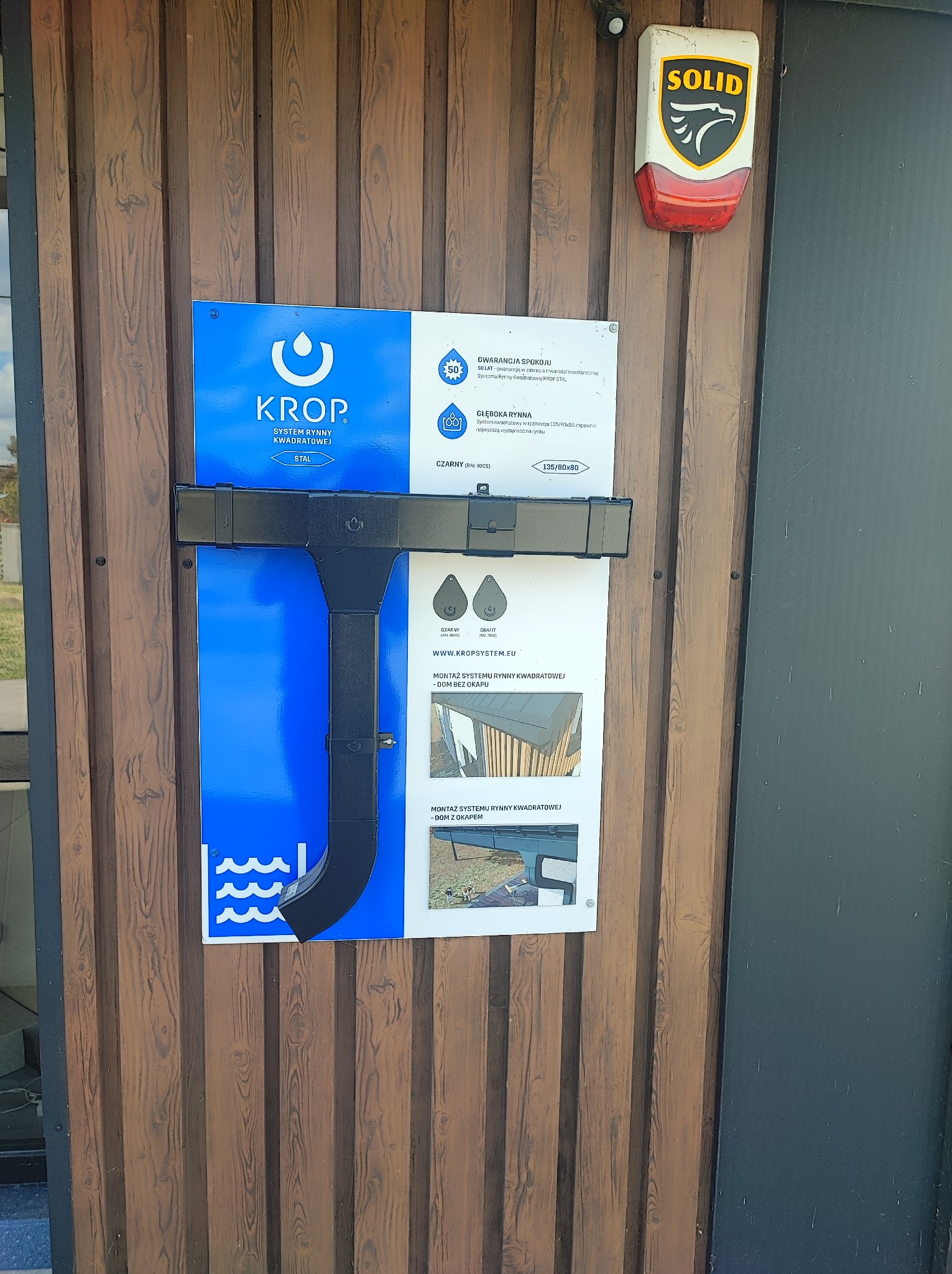 Ekspozycja systemu rynnowego KROP na brązowej ścianie. Prezentacja elementów, w tym rynny kwadratowej, z informacjami o gwarancji i montażu. Widoczny alarm SOLID.