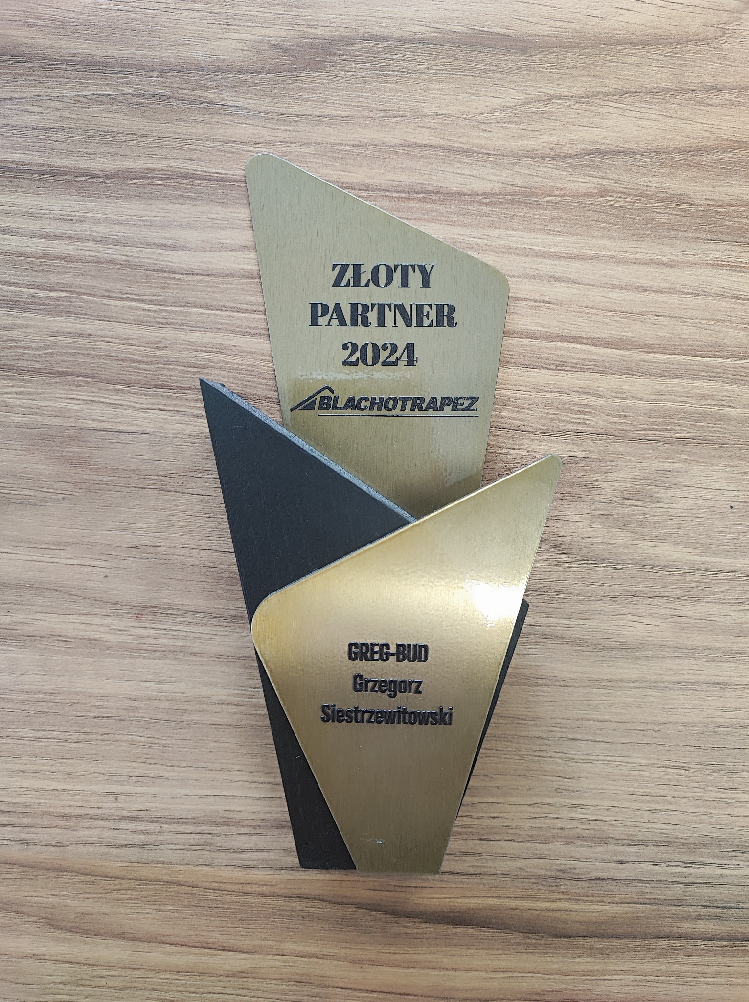 Nagroda 'Złoty Partner 2024' dla firmy BLACHOTRAPEZ oraz Greg-Bud Grzegorz Siestrzewitowski, na drewnianym tle. Trapezoidalny kształt, złote i czarne elementy.