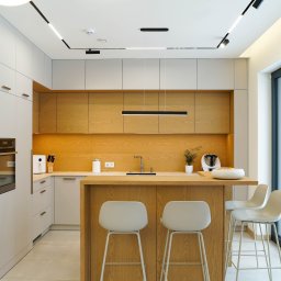 Hubertus Complex - Nowoczesna kuchnia z drewnianymi akcentami: jasne szafki, drewniane blaty i wyspa z trzema białymi hokerami. Duże okno z widokiem na zaśnieżony ogród.