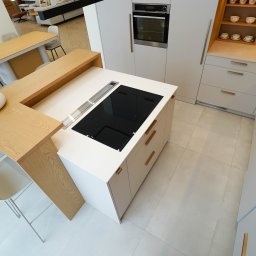 Hubertus Complex - Nowoczesna, jasna kuchnia z wyspą i blatem z naturalnego drewna. Na blacie indukcja i owoce. Minimalistyczne krzesła i stół w tle. Widok z góry.