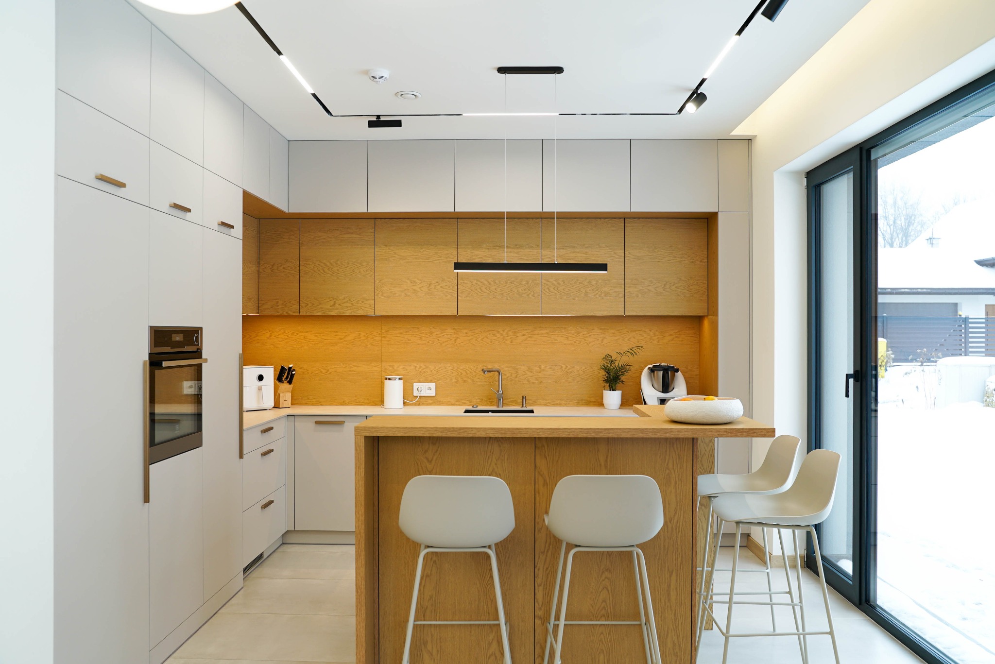 Nowoczesna kuchnia z drewnianymi akcentami: jasne szafki, drewniane blaty i wyspa z trzema białymi hokerami. Duże okno z widokiem na zaśnieżony ogród.