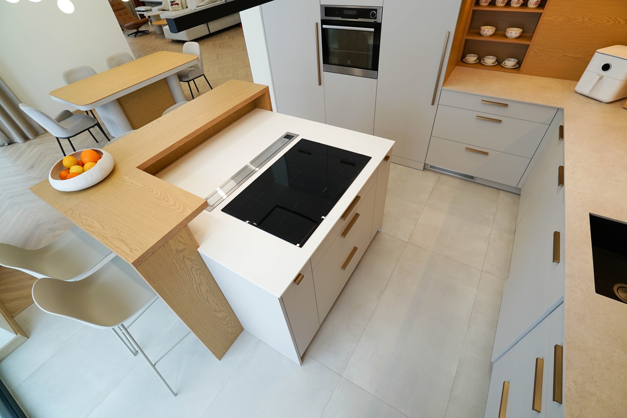 Nowoczesna, jasna kuchnia z wyspą i blatem z naturalnego drewna. Na blacie indukcja i owoce. Minimalistyczne krzesła i stół w tle. Widok z góry.