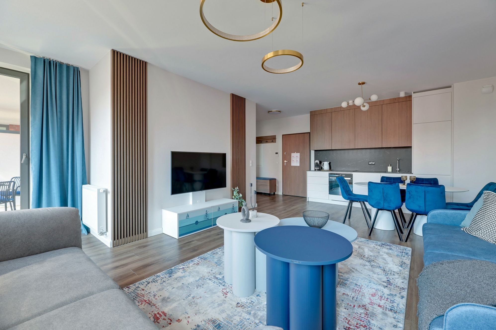 Nowoczesna aranżacja wnętrza salonu z aneksem kuchennym w Siemianowicach Śląskich. Uwagę przykuwają okrągłe stoliki kawowe i drewniane lamele na ścianie.