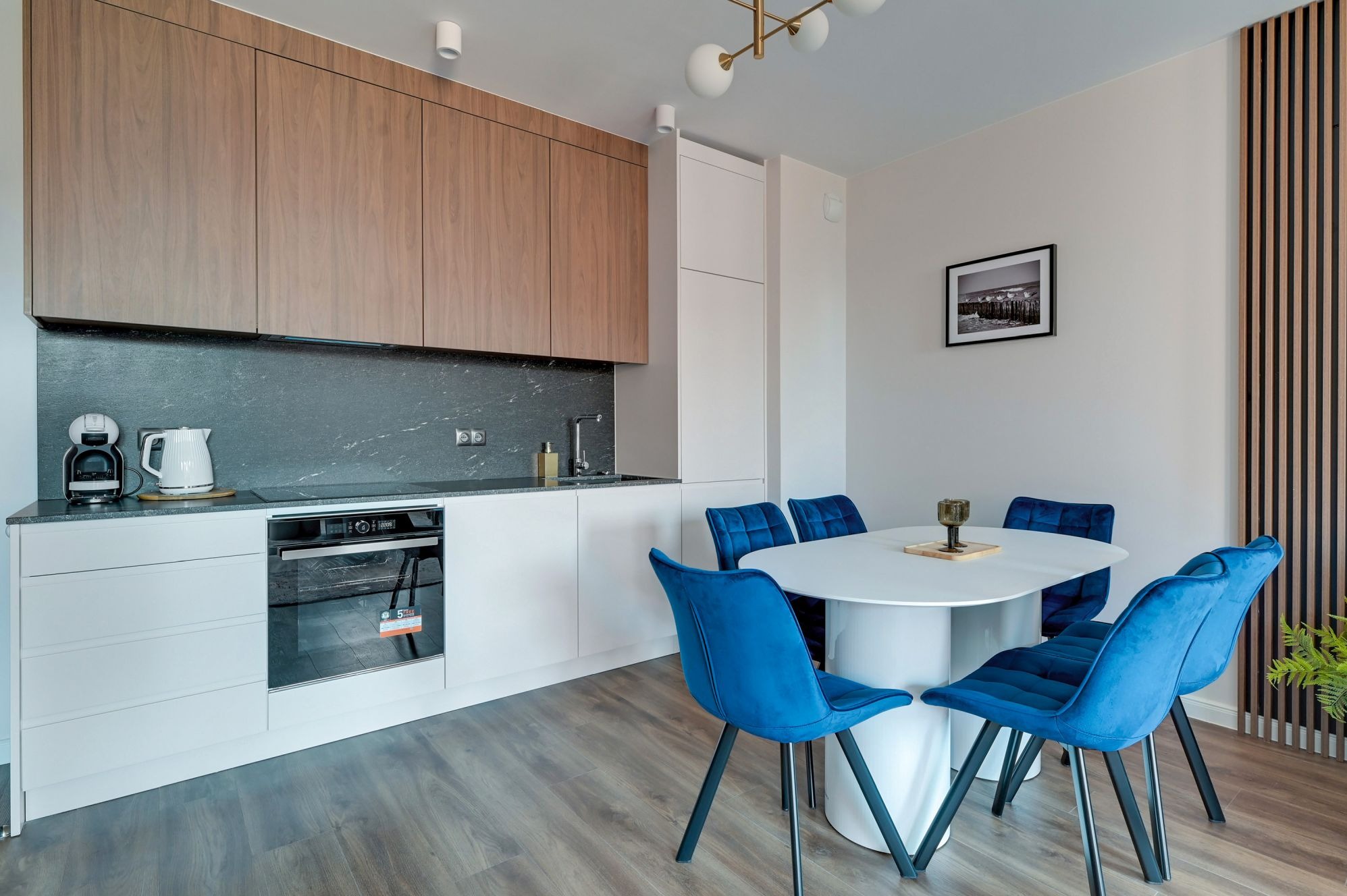 Nowoczesna kuchnia z drewnianymi szafkami górnymi i białymi dolnymi, uzupełniona okrągłym stołem i welurowymi, niebieskimi krzesłami. Minimalistyczny design.