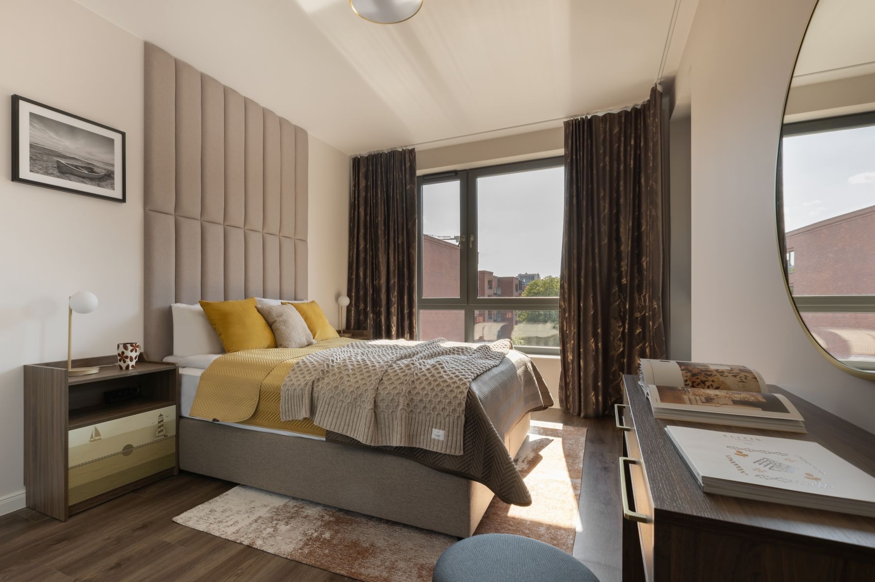 Nowoczesna sypialnia: łóżko z tapicerowanym zagłówkiem, stolik nocny z motywem marynistycznym, brązowe zasłony i lustro w złotej ramie. Widok na miasto przez okno.