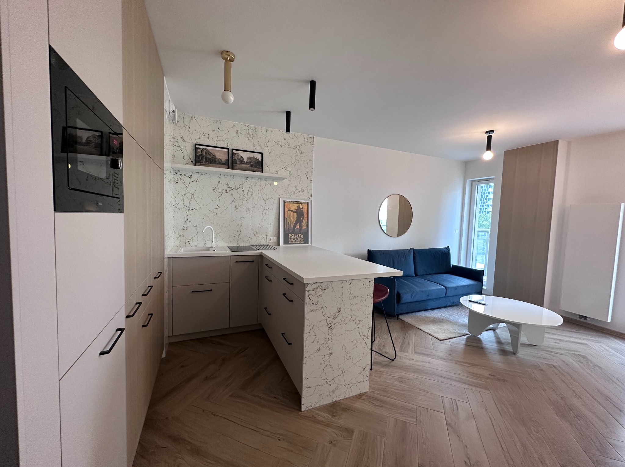 Jasna, nowoczesna kuchnia z wyspą i marmurowym akcentem, połączona z salonem. Parkiet w jodełkę, minimalistyczne oświetlenie, niebieska sofa w tle.