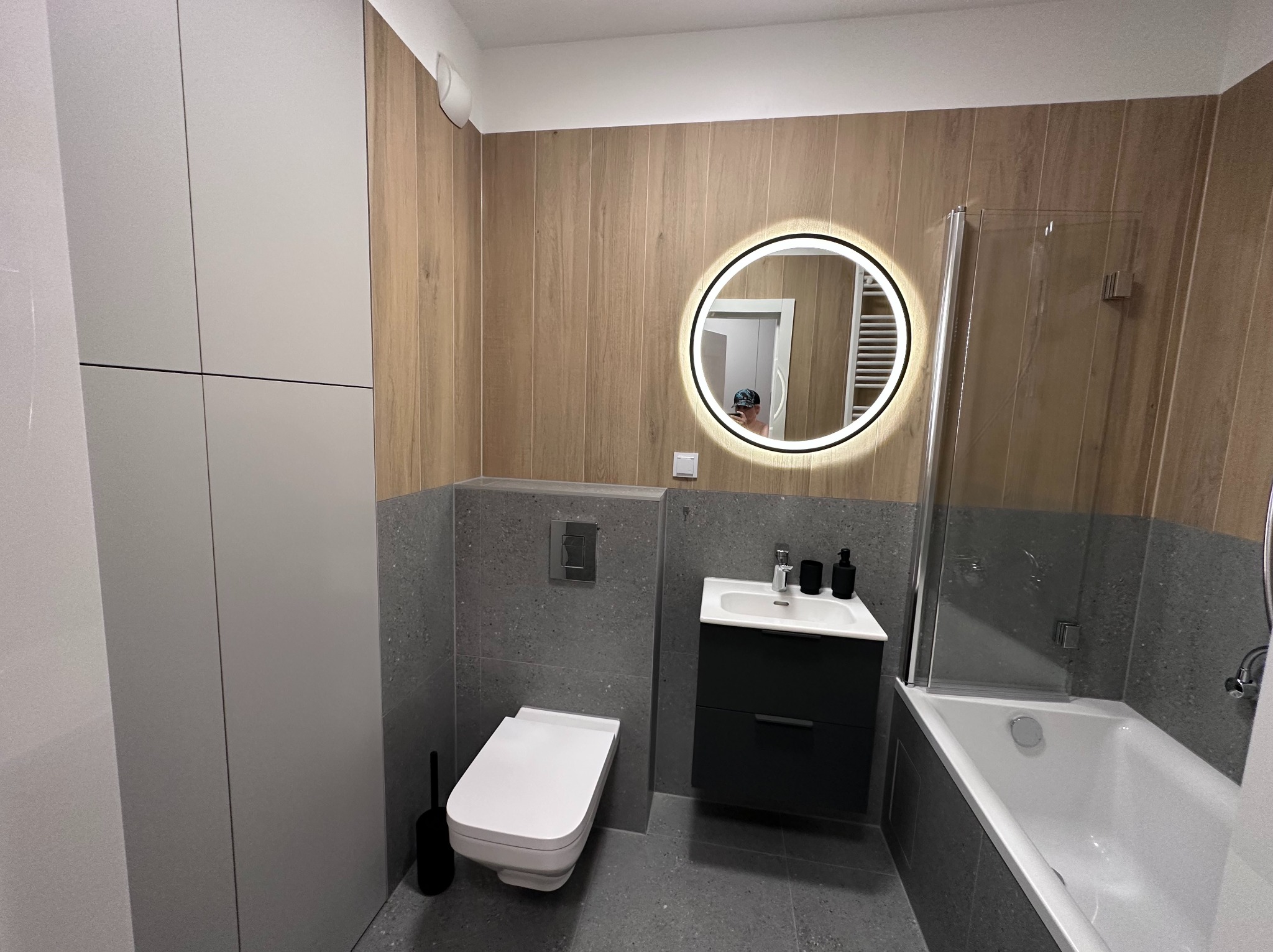Nowoczesna łazienka z drewnopodobnymi płytkami, szarą strefą umywalki i toalety, okrągłym lustrem z podświetleniem i zabudową meblową w jasnym odcieniu szarości.