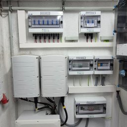 Kacper Kosmala - Rozdzielnice DC i AC fotowoltaiki, inwertery zamontowane na ścianie w pomieszczeniu technicznym. Widoczne przewody i okablowanie instalacji.