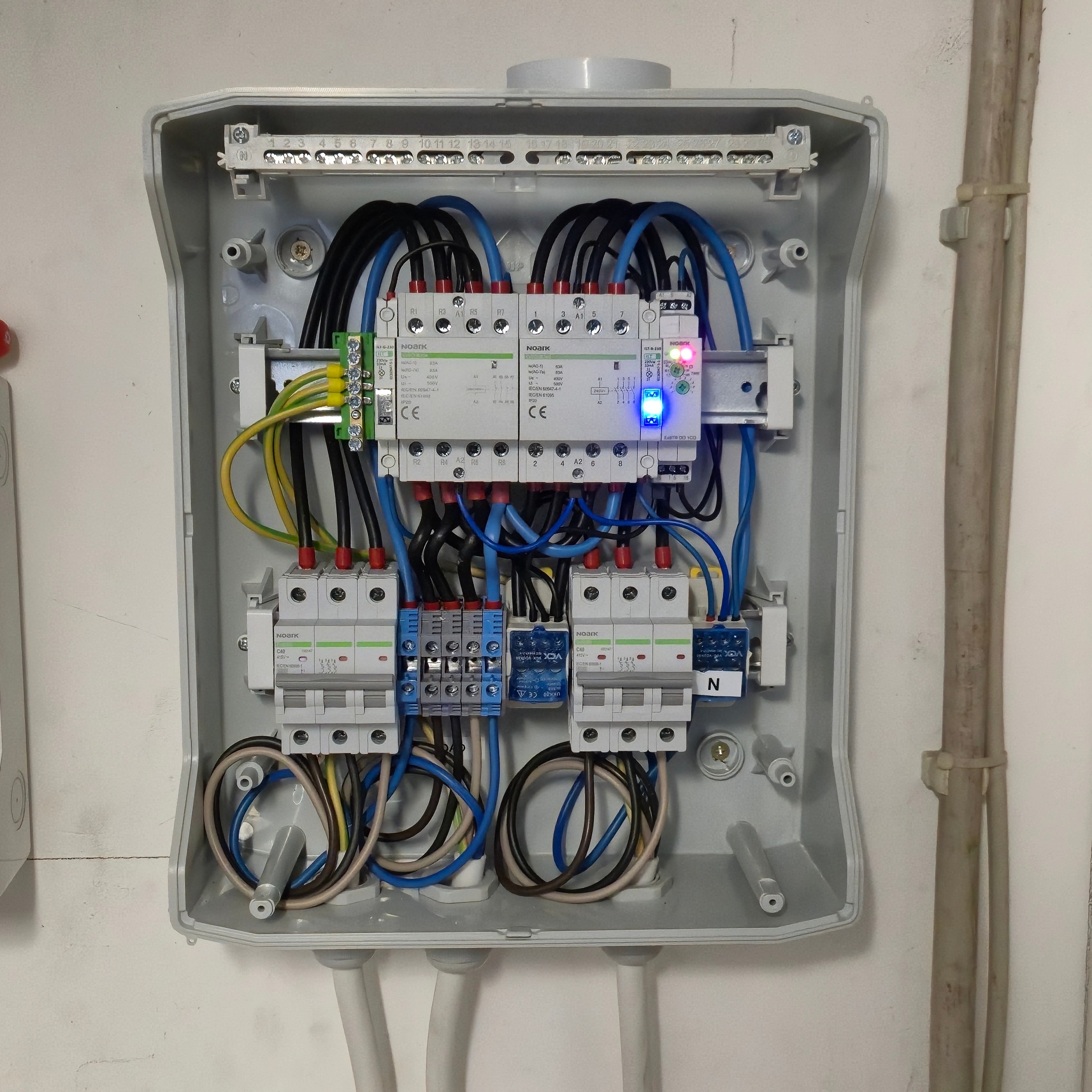 Szara skrzynka rozdzielcza z zainstalowanymi bezpiecznikami, przewodami i modułami. Widoczne są kable w kolorach czarnym, niebieskim i żółto-zielonym. Elementy są marki Noark.