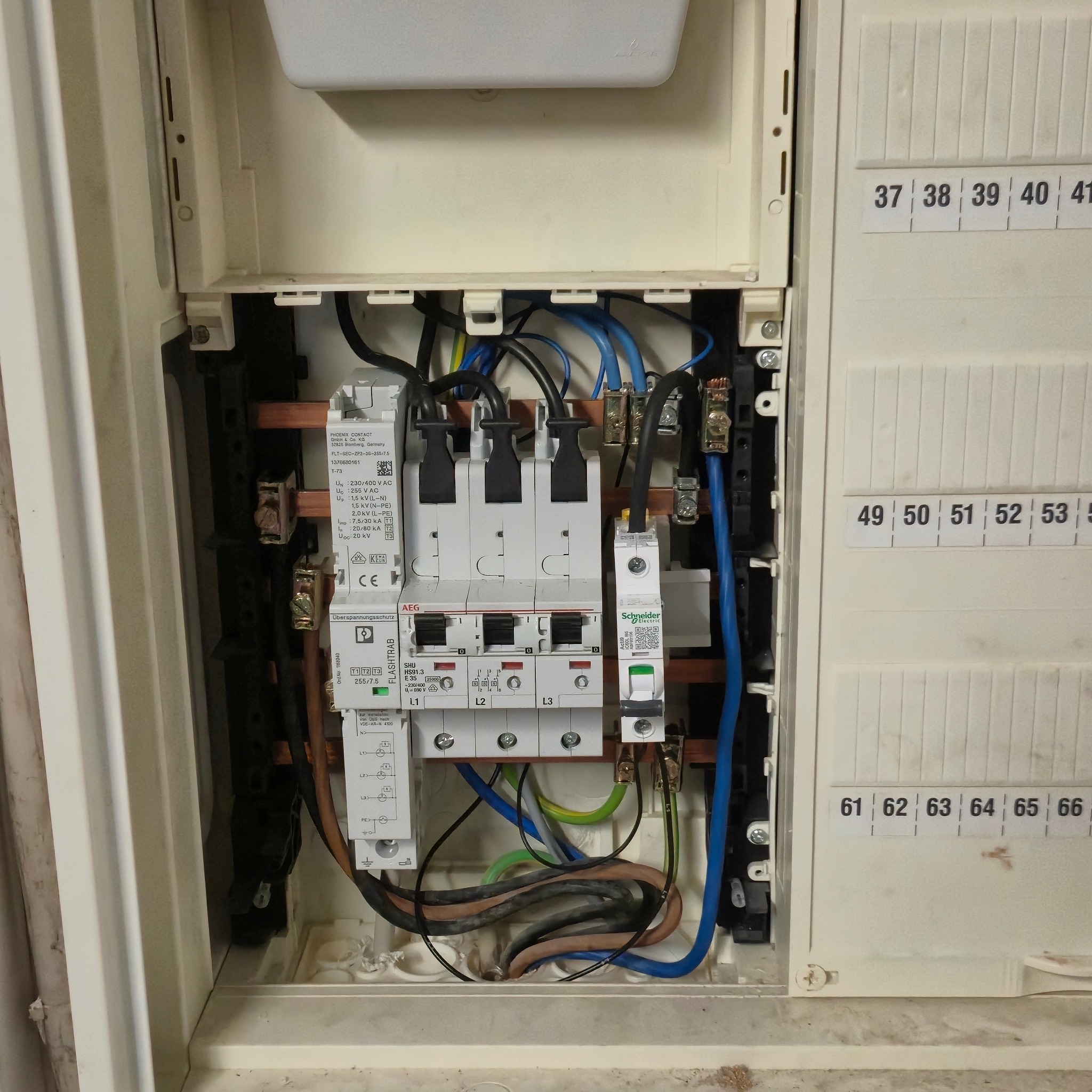 Otwarta skrzynka bezpiecznikowa z widocznymi bezpiecznikami, przewodami i listwami. Tabliczka z numeracją obwodów po prawej stronie. Widoczne elementy instalacji elektrycznej.