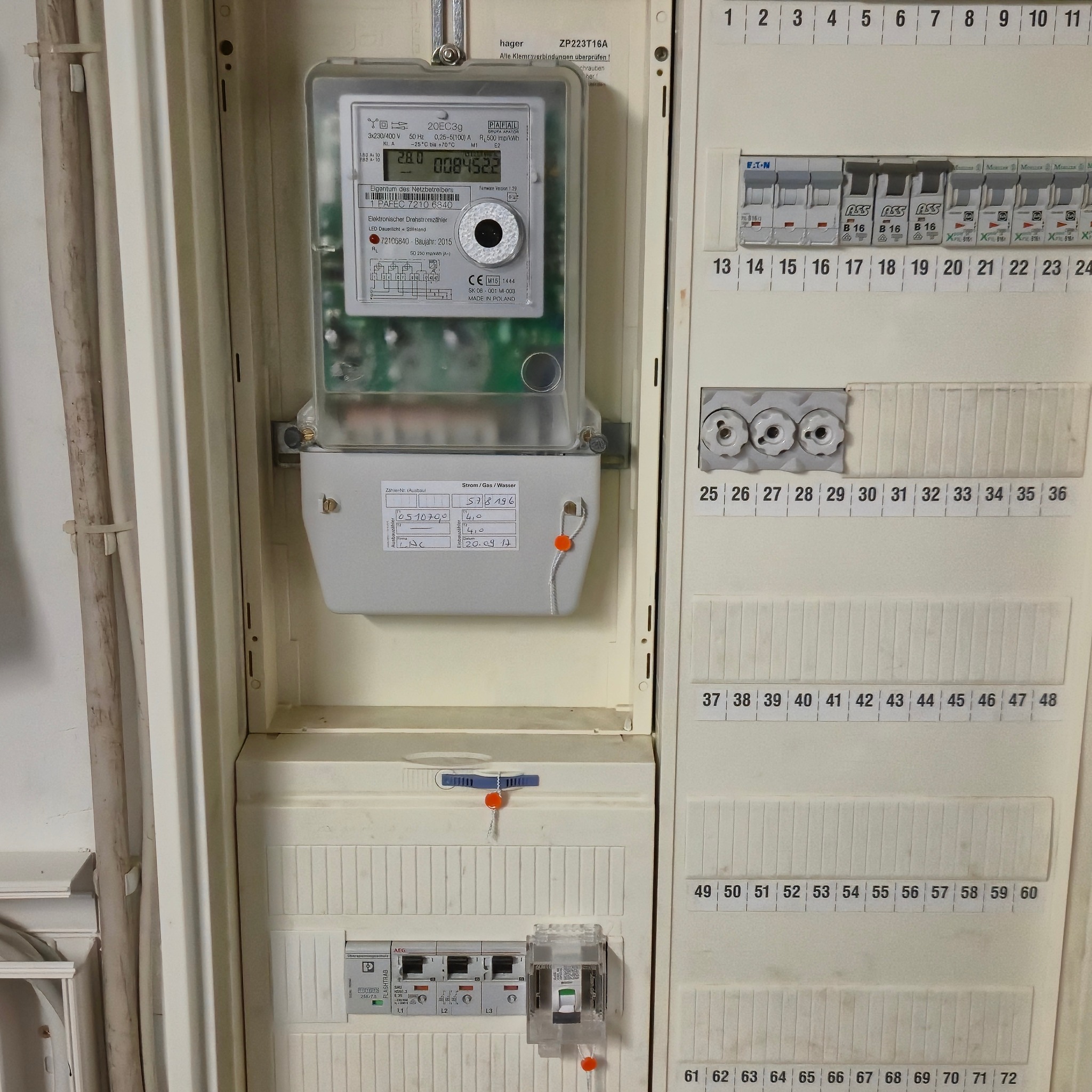 Szczegółowe ujęcie skrzynki elektrycznej z licznikiem energii, bezpiecznikami i oznaczeniami numerów. Widoczne elementy instalacji elektrycznej wewnątrz budynku.