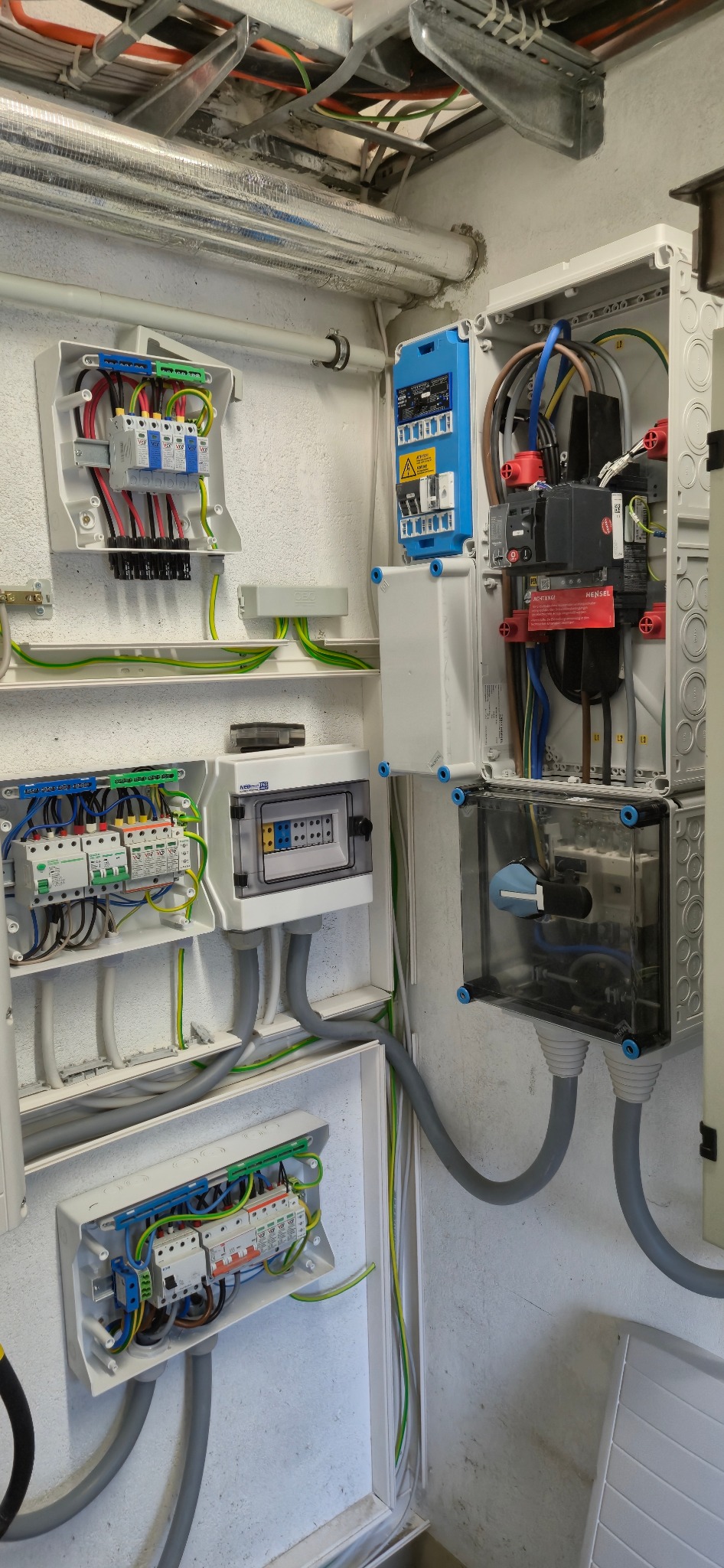 Rozdzielnice elektryczne w białych obudowach, z widocznymi przewodami, bezpiecznikami i elementami instalacji, zamontowane na ścianie wewnątrz budynku.