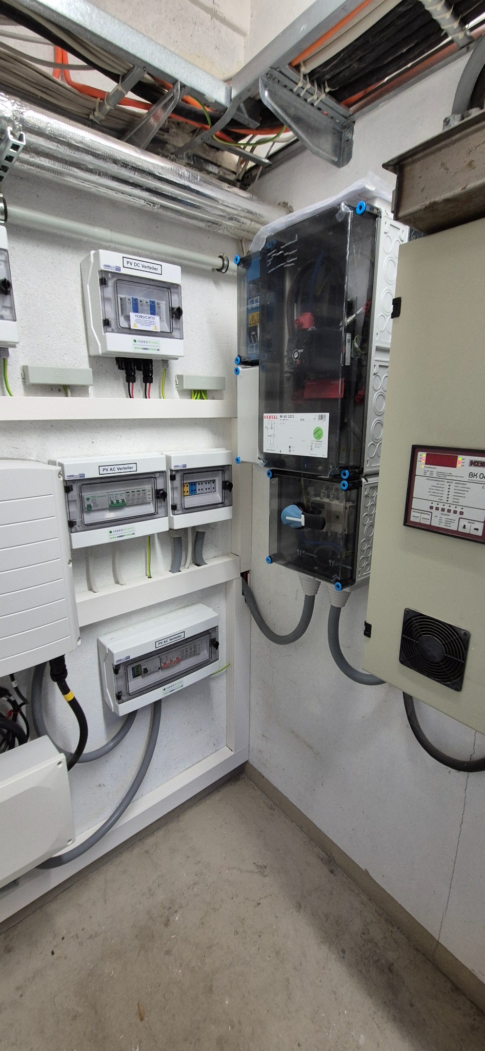 Rozdzielnice PV DC i AC w białych obudowach, zamontowane na ścianie. Widoczne kable i rury instalacyjne. Górą metalowe korytka kablowe.