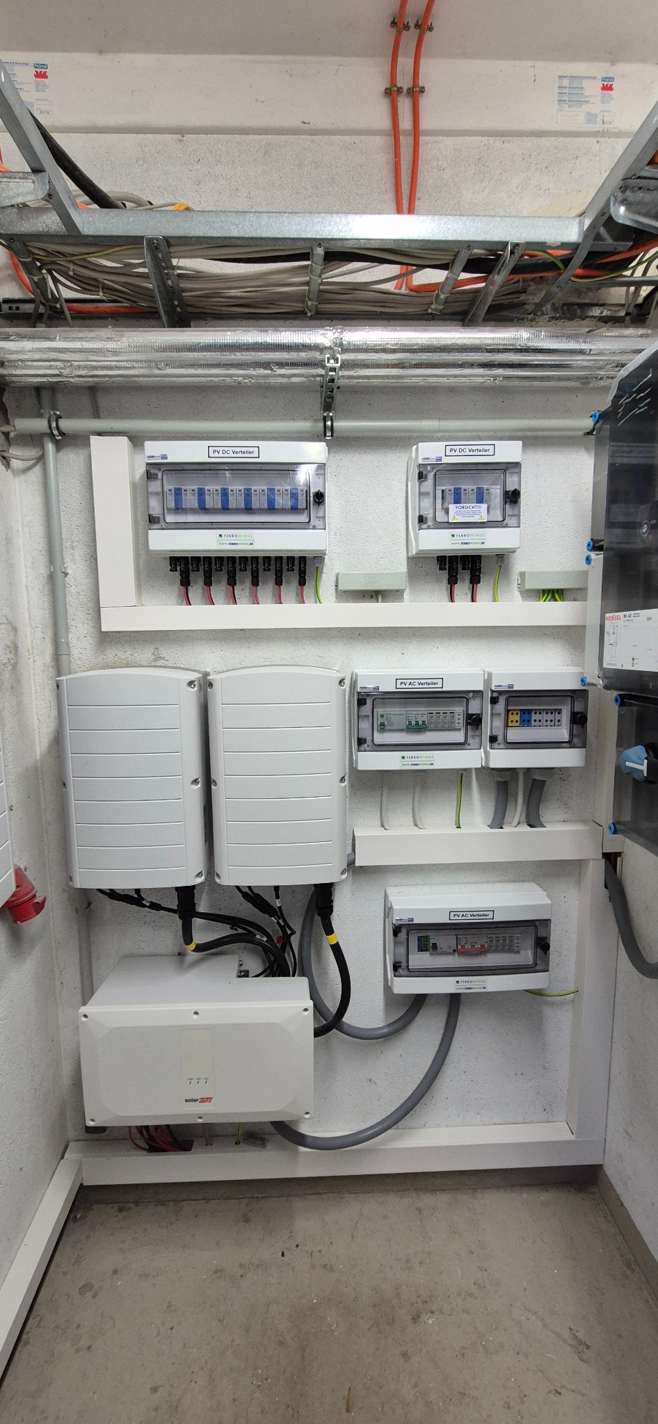Rozdzielnice DC i AC fotowoltaiki, inwertery zamontowane na ścianie w pomieszczeniu technicznym. Widoczne przewody i okablowanie instalacji.
